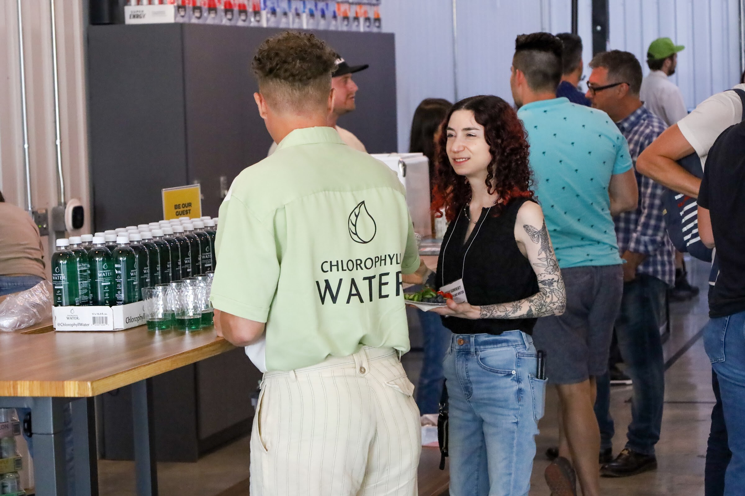 A man in a light green Chlorophyll Water bowling shirt with his back to the camera talks with a woman with curly red hair holding a plate of food, beside a wooden table with a case of green Chlorophyll Water bottles and cups.