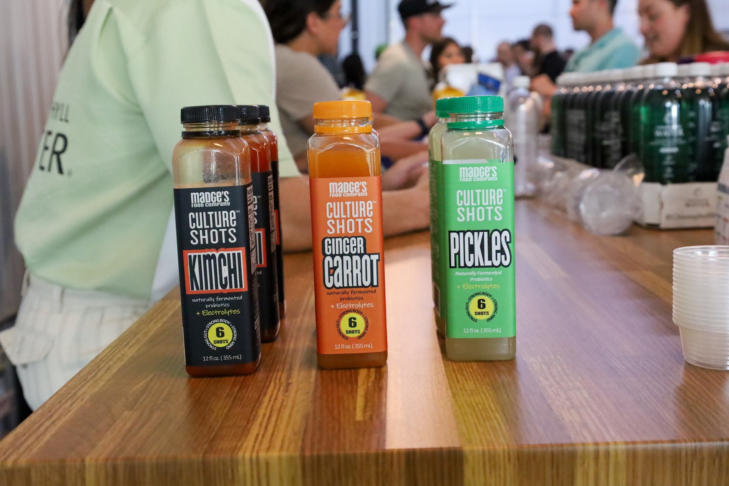 Bottles of Madge's Food Company Culture Shots in Kimchi, Ginger Carrot and Pickles flavors stand on a wooden table, with a case of Chlorophyll Water bottles and a stack of plastic cups behind them.