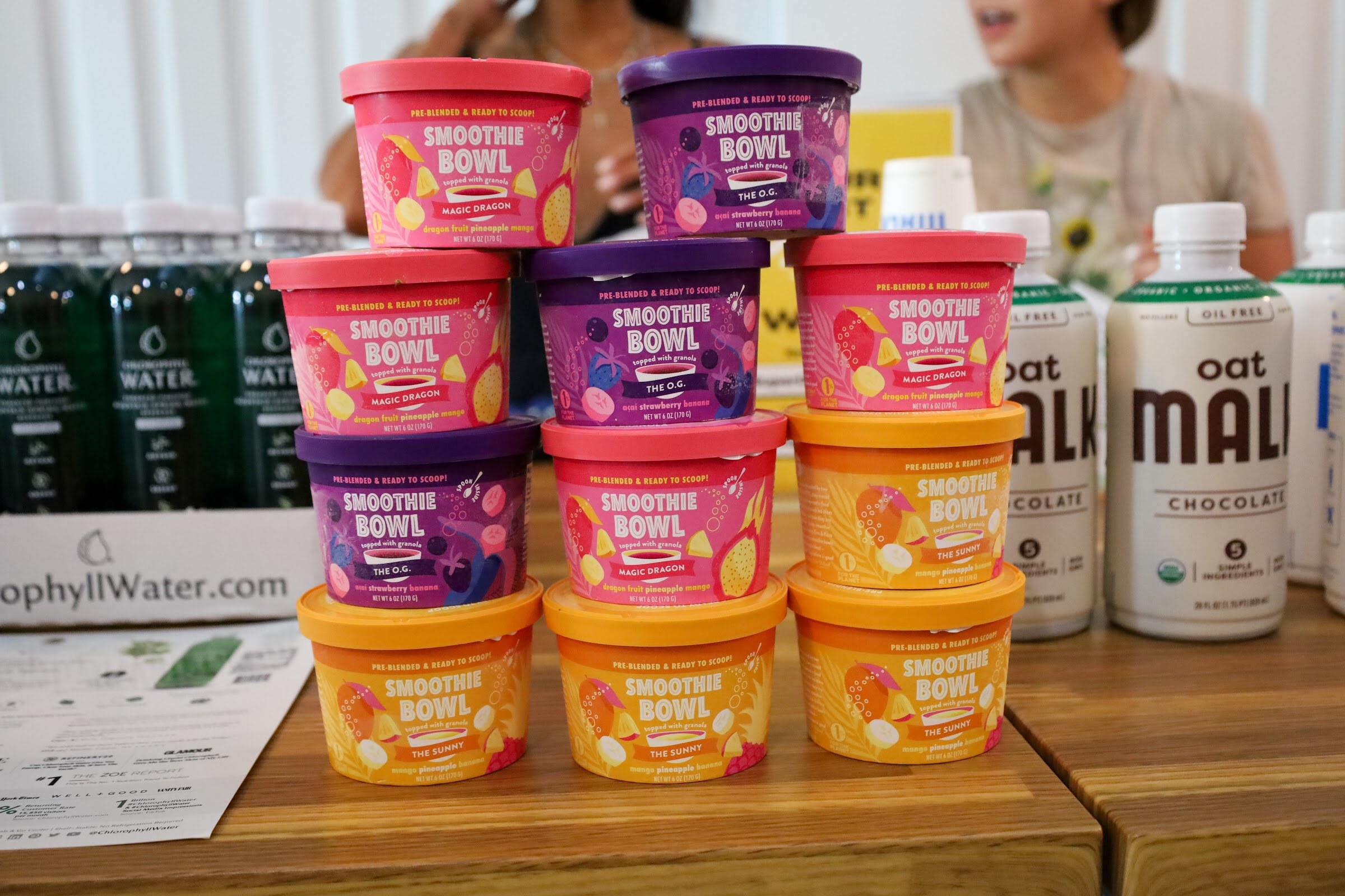 Stacks of Smoothie Bowl cups in Magic Dragon, The O.G. and The Sunny flavors beside bottles of oat MALK Chocolate and Chlorophyll Water on a wooden table.