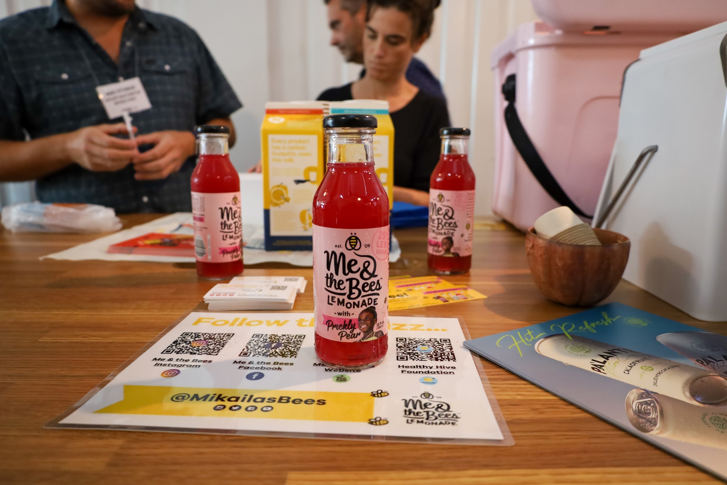 Three bottles of Me & the Bees Lemonade with Prickly Pear stand on a wooden table in front of a sign with QR codes and the handle @MikailasBees, with a Palante can at the right and two people seated behind the table.