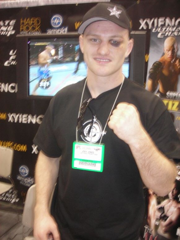Joey Jones, in a black cap and shirt with a bruised eye, raises a fist in front of Xyience, UFC, Hard Rock Hotel & Casino and ampd mobile signs and a screen showing a fight.