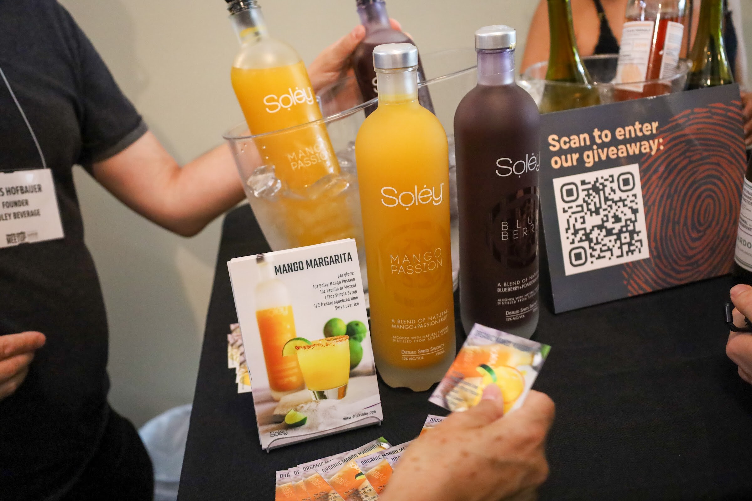 Bottles of Soley Mango Passion and Blueberry stand beside an ice bucket on a black table with a Mango Margarita recipe card and a sign reading Scan to enter our giveaway, with a person wearing a name badge at the left.