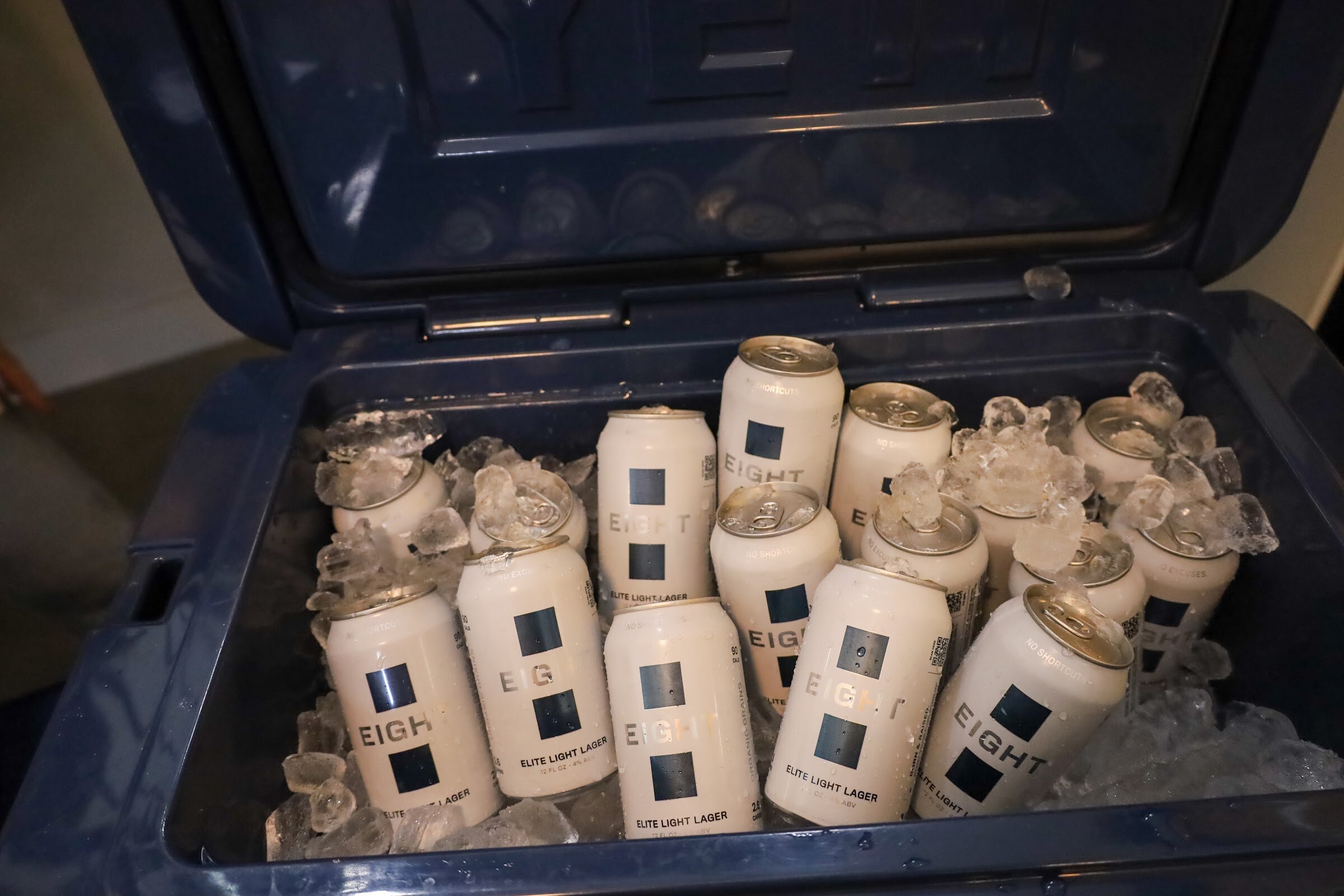 White cans of EIGHT Elite Light Lager in ice inside an open dark blue cooler.