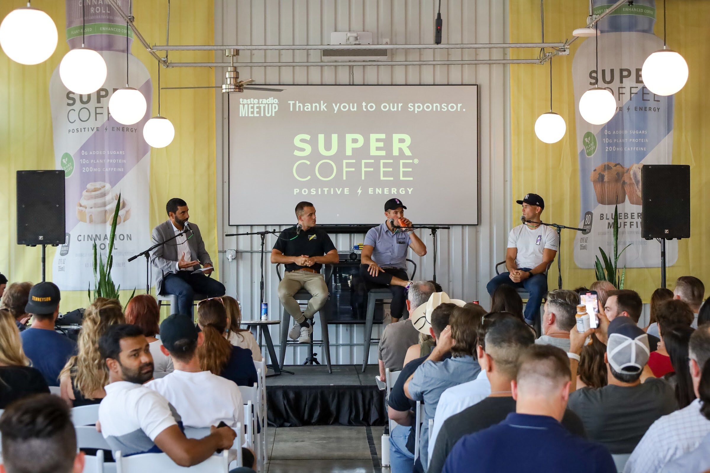 A man in a gray blazer holding notes and three men in a black polo, a blue-gray polo and a white t-shirt and cap sit on gray bar stools with microphones in front of a screen reading Taste Radio Meetup, Thank you to our sponsor, Super Coffee, with a seated audience in the foreground.