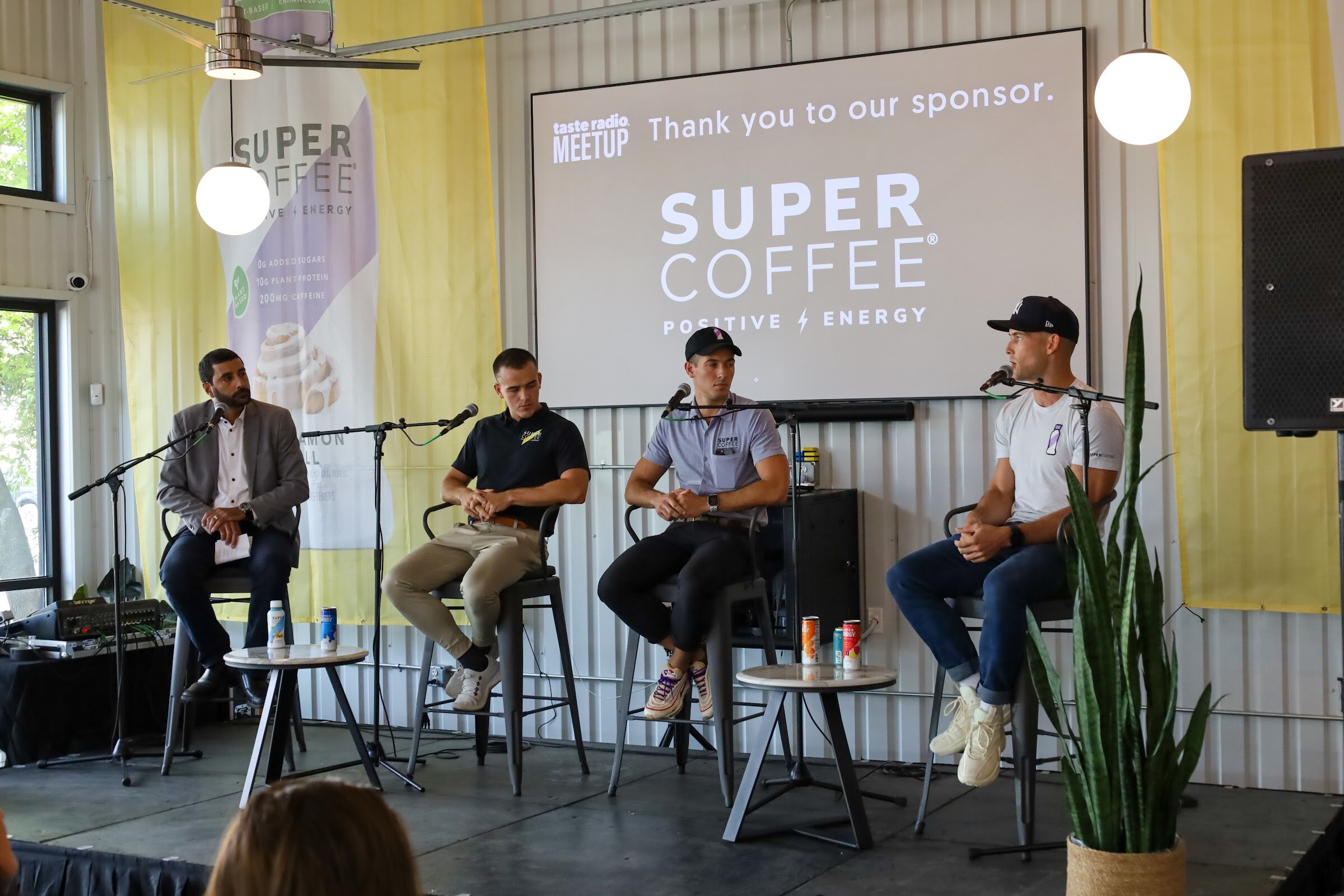 A man in a gray blazer, a man in a black polo, a man in a blue-gray polo and black cap and a man in a white t-shirt and black cap sit on tall stools with microphones in front of the sponsor screen, beside a tall plant, with cans on small tables.