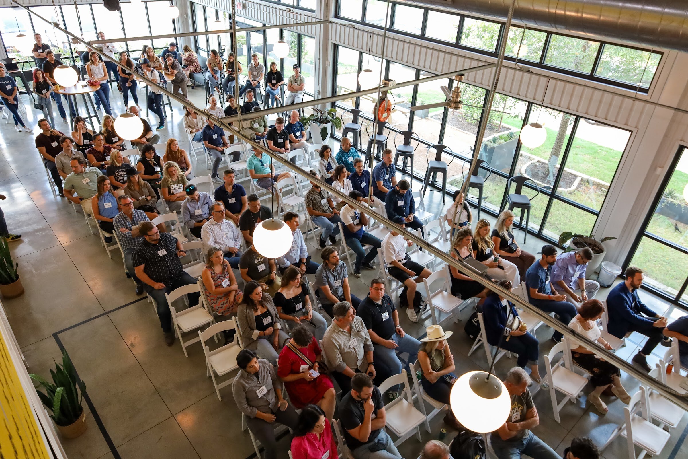 High-angle view of seated attendees with name badges in white folding chairs on a concrete floor in a bright warehouse-style room with tall windows and round pendant lights, with bar stools along the glass.