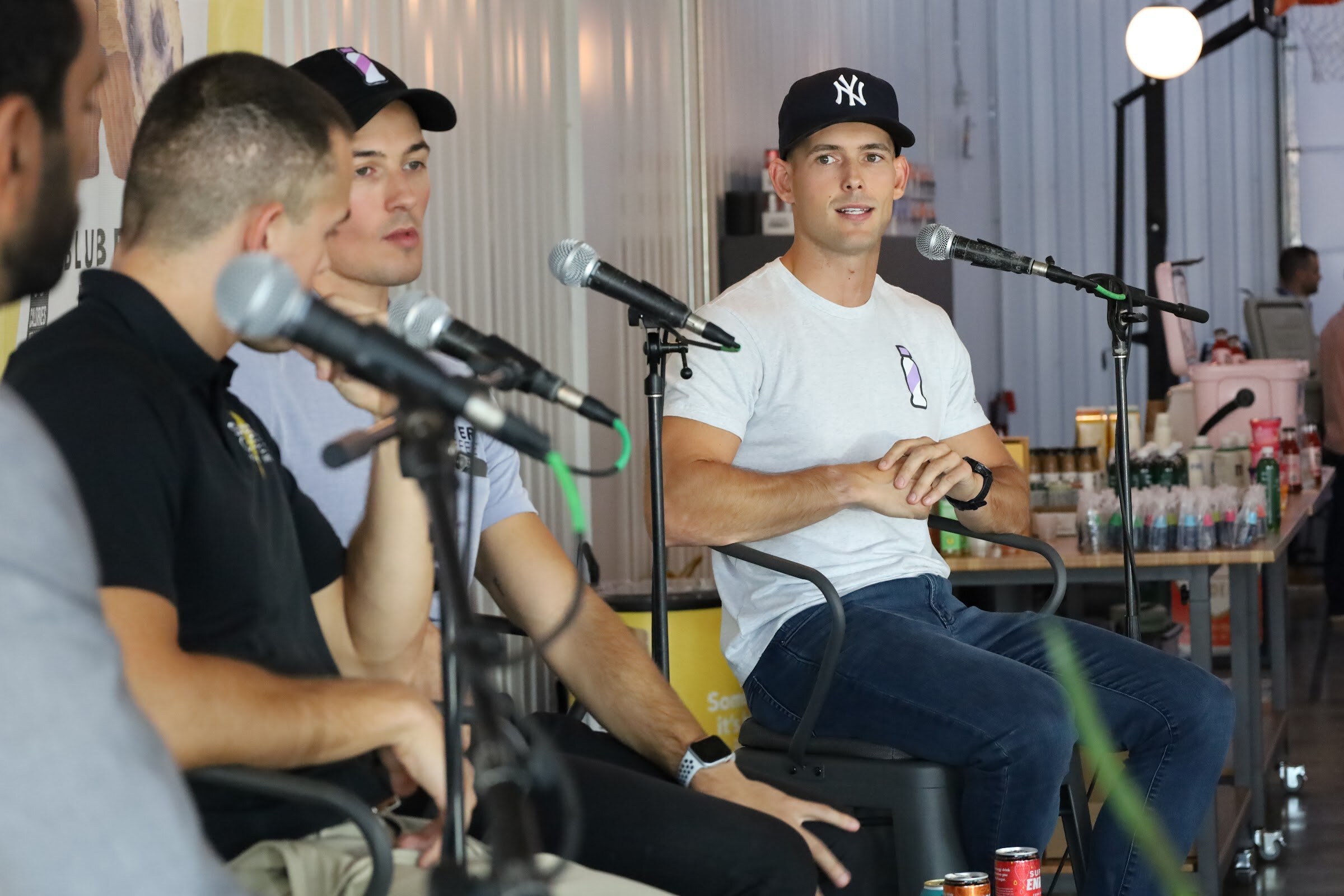 A man in a New York Yankees cap and white t-shirt speaks to the host with his hands clasped, with microphones on stands, two other guests in the foreground and a sampling table with bottles behind him.