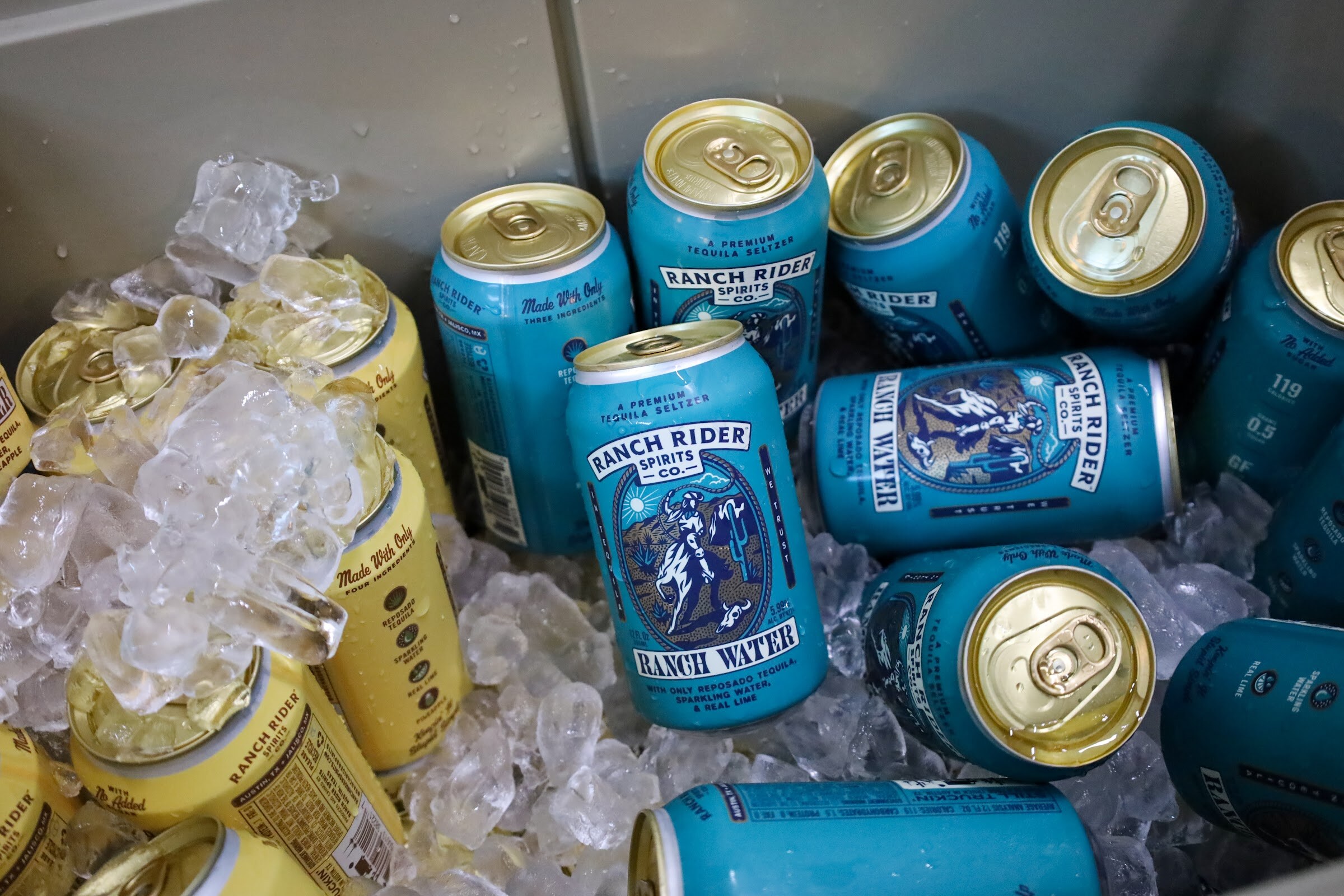 Teal Ranch Rider Spirits Co. Ranch Water cans and yellow cans lie in ice in a gray bin.