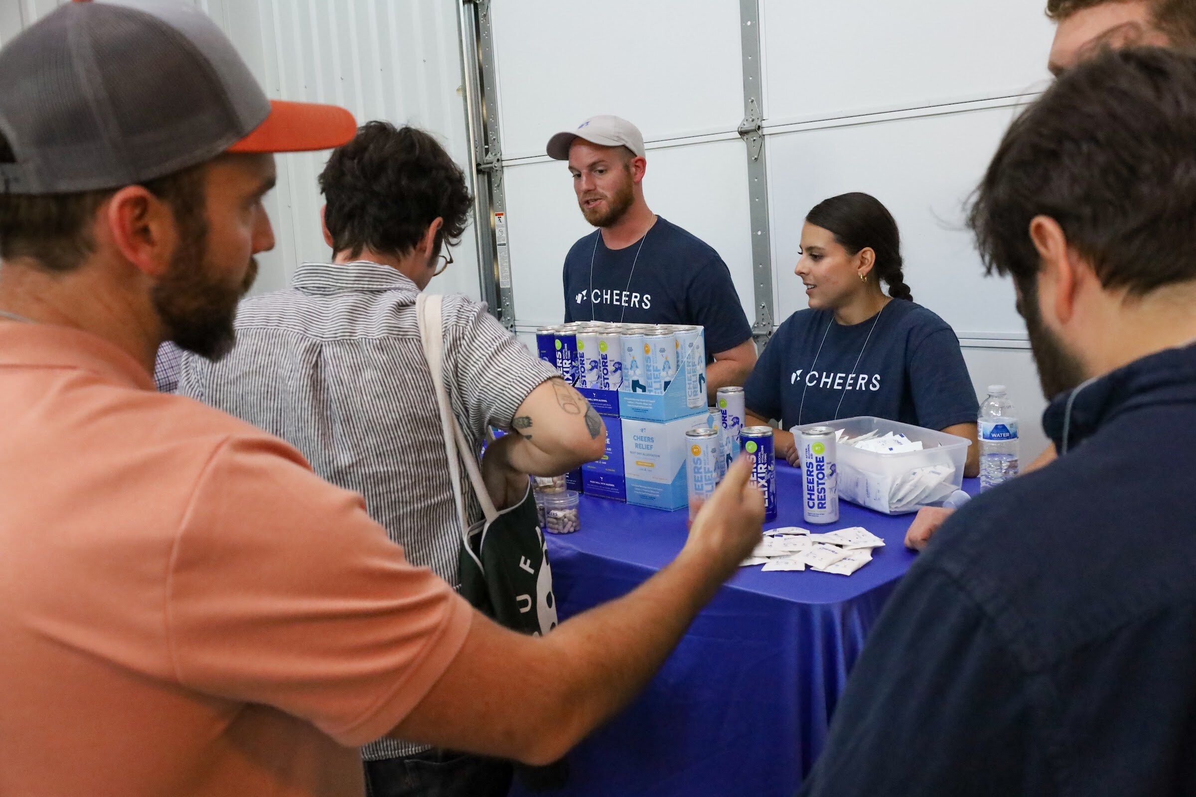 A man and a woman in navy Cheers t-shirts stand behind a blue-draped table with cans, boxes and a bin of packets, while guests in the foreground lean in.