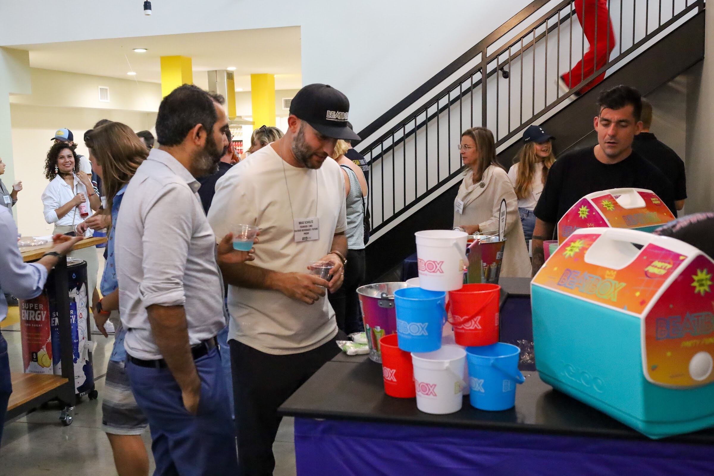 A man in a black Beatbox cap and cream t-shirt holds a small cup beside a table of Beatbox buckets and a turquoise Beatbox Party Punch cooler, with other guests, a Super Enrgy display and a staircase behind.