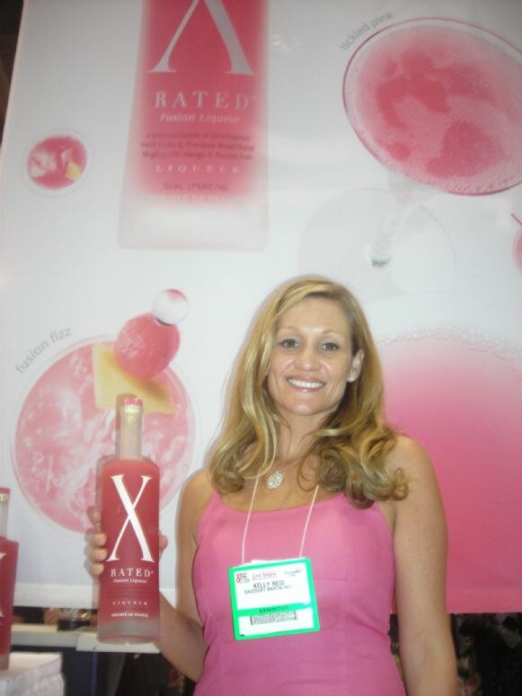 Kelly Reid, wearing a pink top and an exhibitor badge, smiles and holds a bottle of X Rated Fusion Liqueur in front of a pink banner showing the bottle and cocktails.