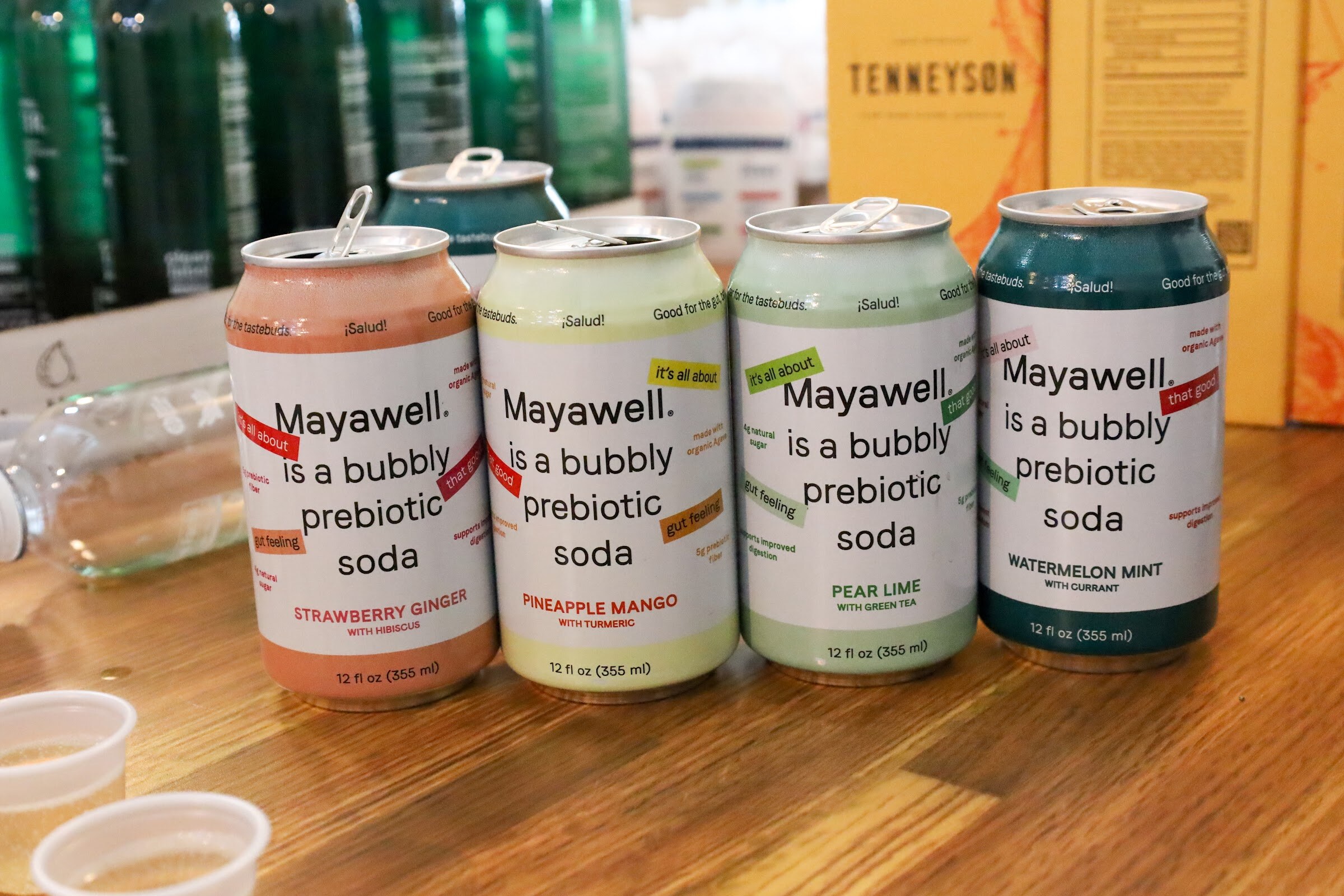 Cans of Mayawell bubbly prebiotic soda in Strawberry Ginger, Pineapple Mango, Pear Lime and Watermelon Mint stand on a wooden table in front of Tenneyson boxes and green bottles.