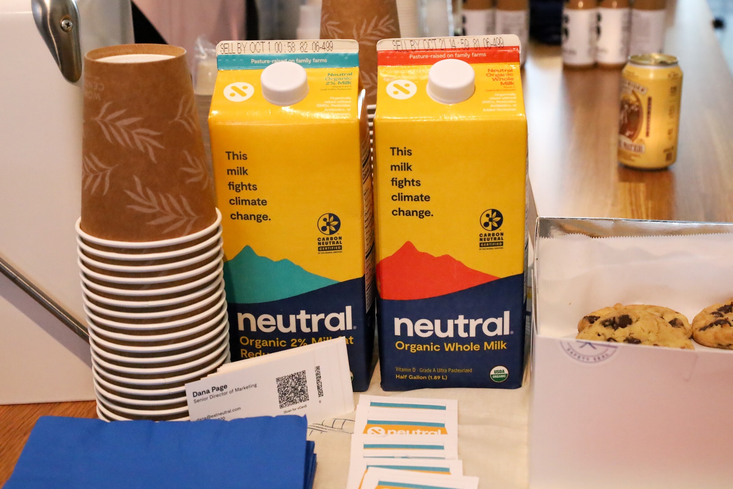 Two cartons of neutral Organic milk labeled This milk fights climate change beside a stack of paper cups, a box of cookies, a blue napkin and business cards.