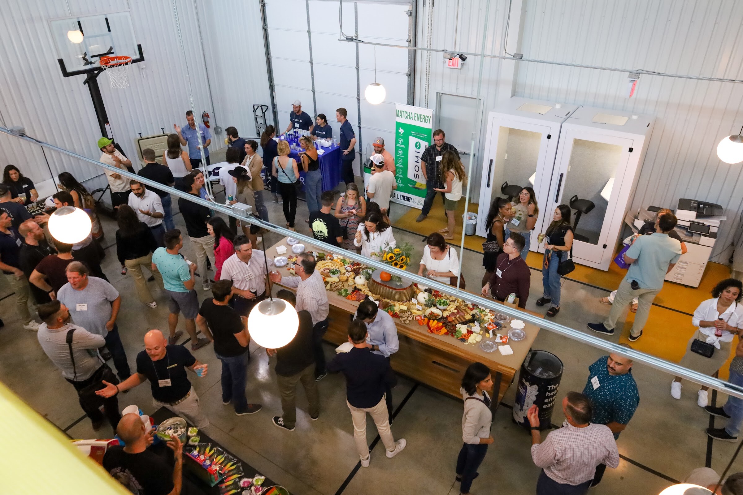 View from above of guests with name badges standing around a long wooden grazing table with a Somi Matcha Energy banner, a blue-draped table and a basketball hoop in the background.