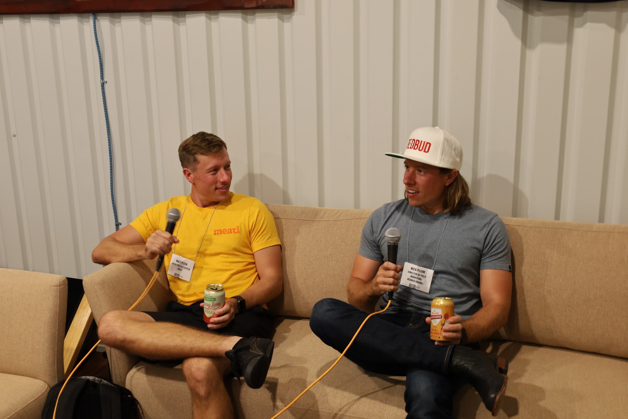 A man in a yellow meati t-shirt and a man in a gray t-shirt and a white Redbud cap sit on a tan sofa holding microphones and cans, both wearing name badges, in front of a corrugated white wall.