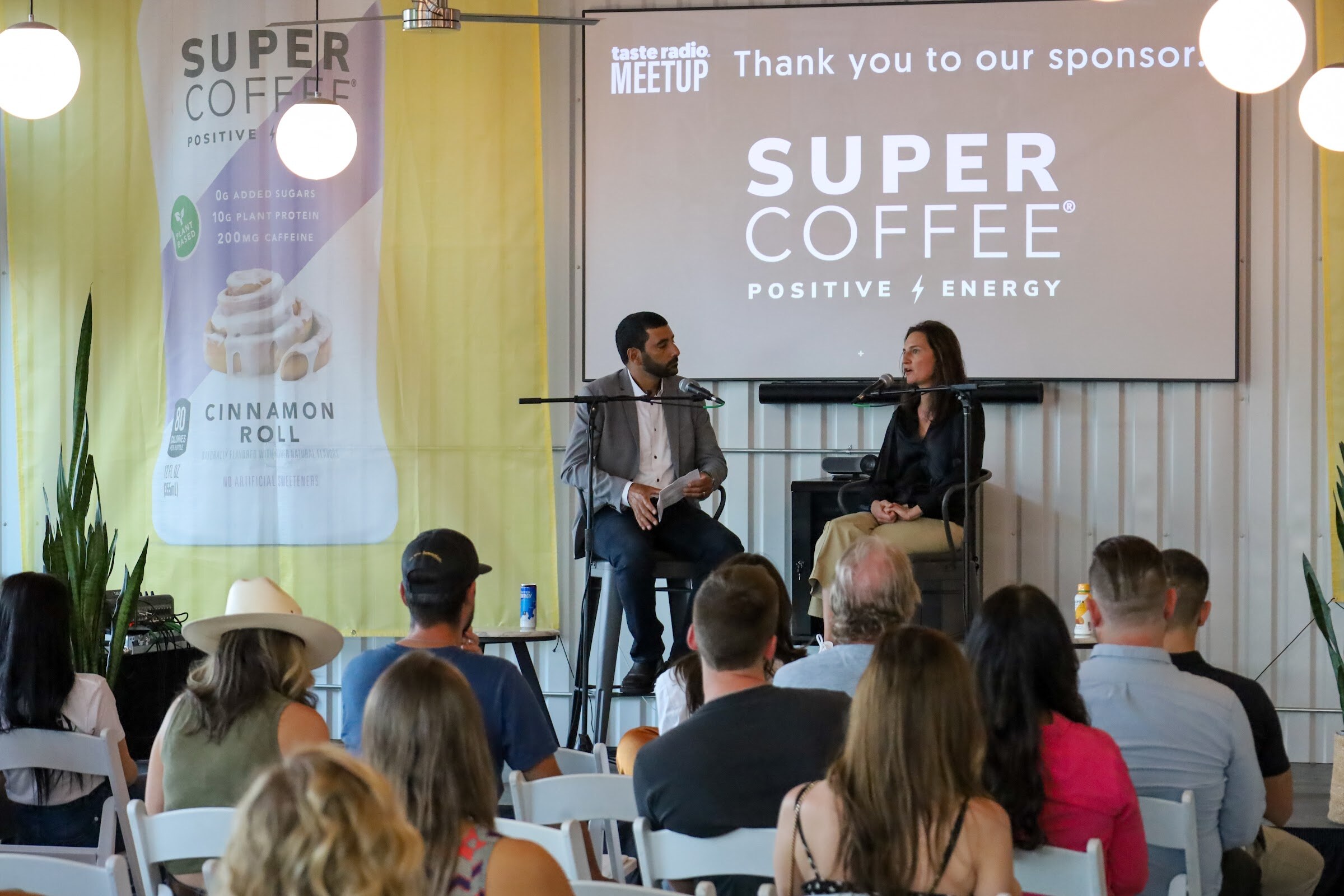 A bearded man in a gray blazer holding notes and a woman in a black satin blouse and khaki pants sit on tall stools with microphones in front of a Super Coffee sponsor screen and a Cinnamon Roll banner, with seated guests in white folding chairs in the foreground.