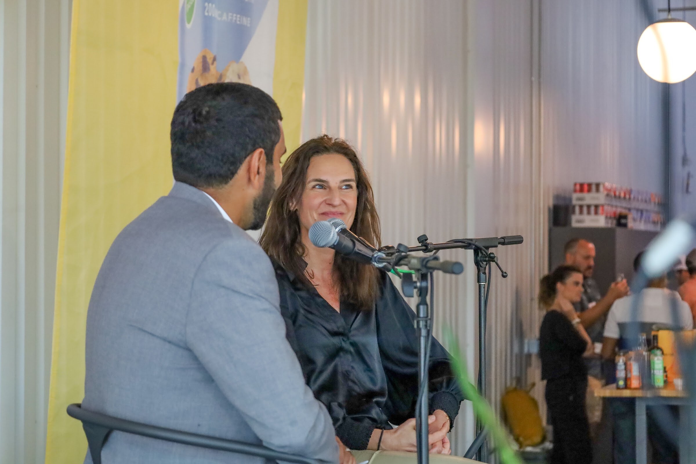 A woman with long brown hair in a black satin blouse smiles toward a man in a gray blazer seen from behind, with a microphone on a stand and guests at a drinks table in the background.
