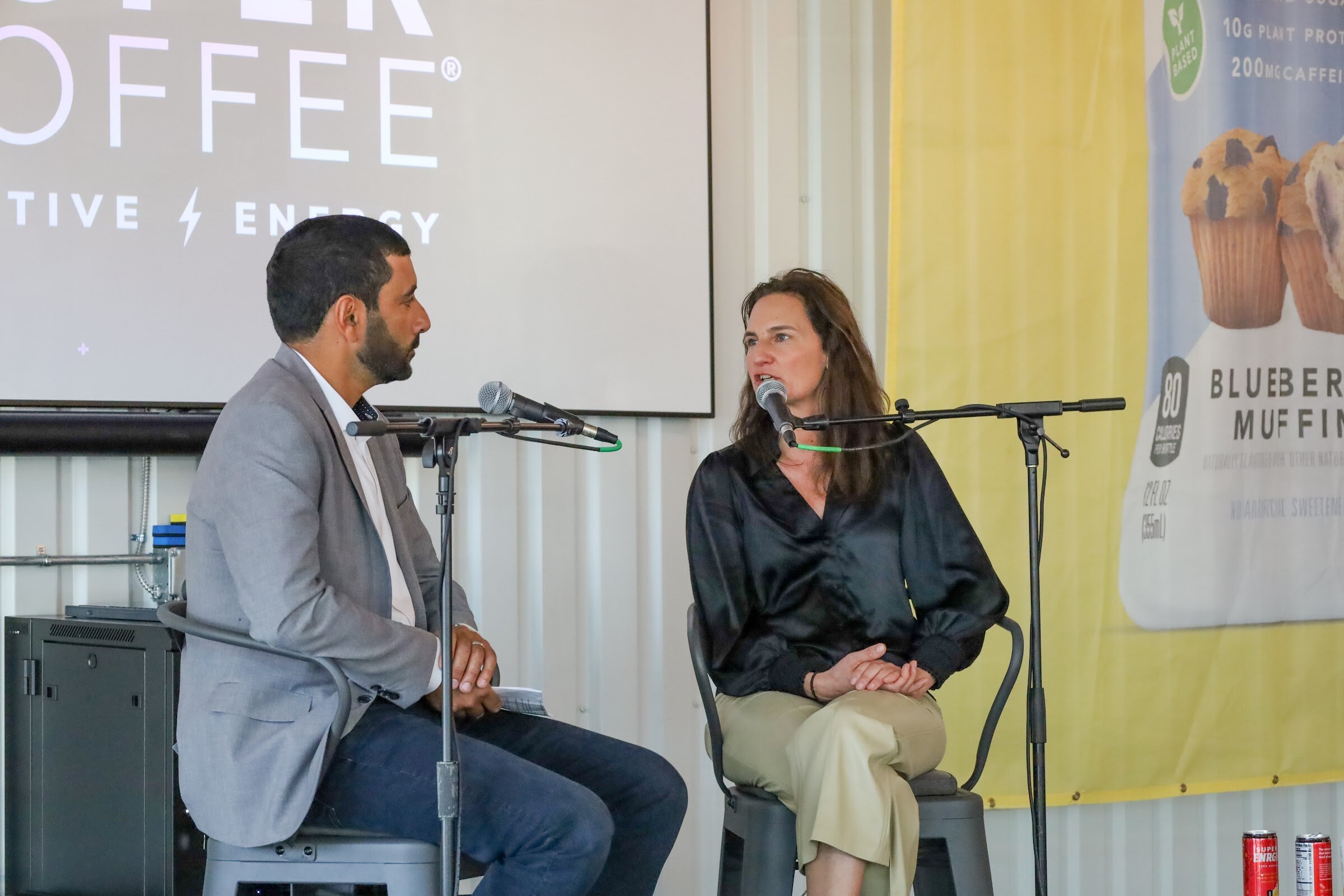 A woman in a black satin blouse and khaki pants speaks into a microphone while a bearded man in a gray blazer listens, in front of a Super Coffee screen and a Blueberry Muffin banner.