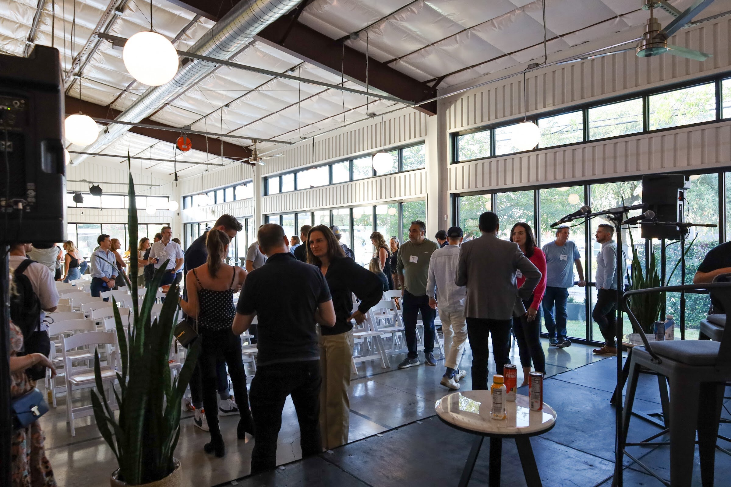 Guests with name badges stand and talk between rows of white folding chairs in a bright warehouse-style room with tall windows, with microphone stands, a small table with cans and a tall plant in the foreground.