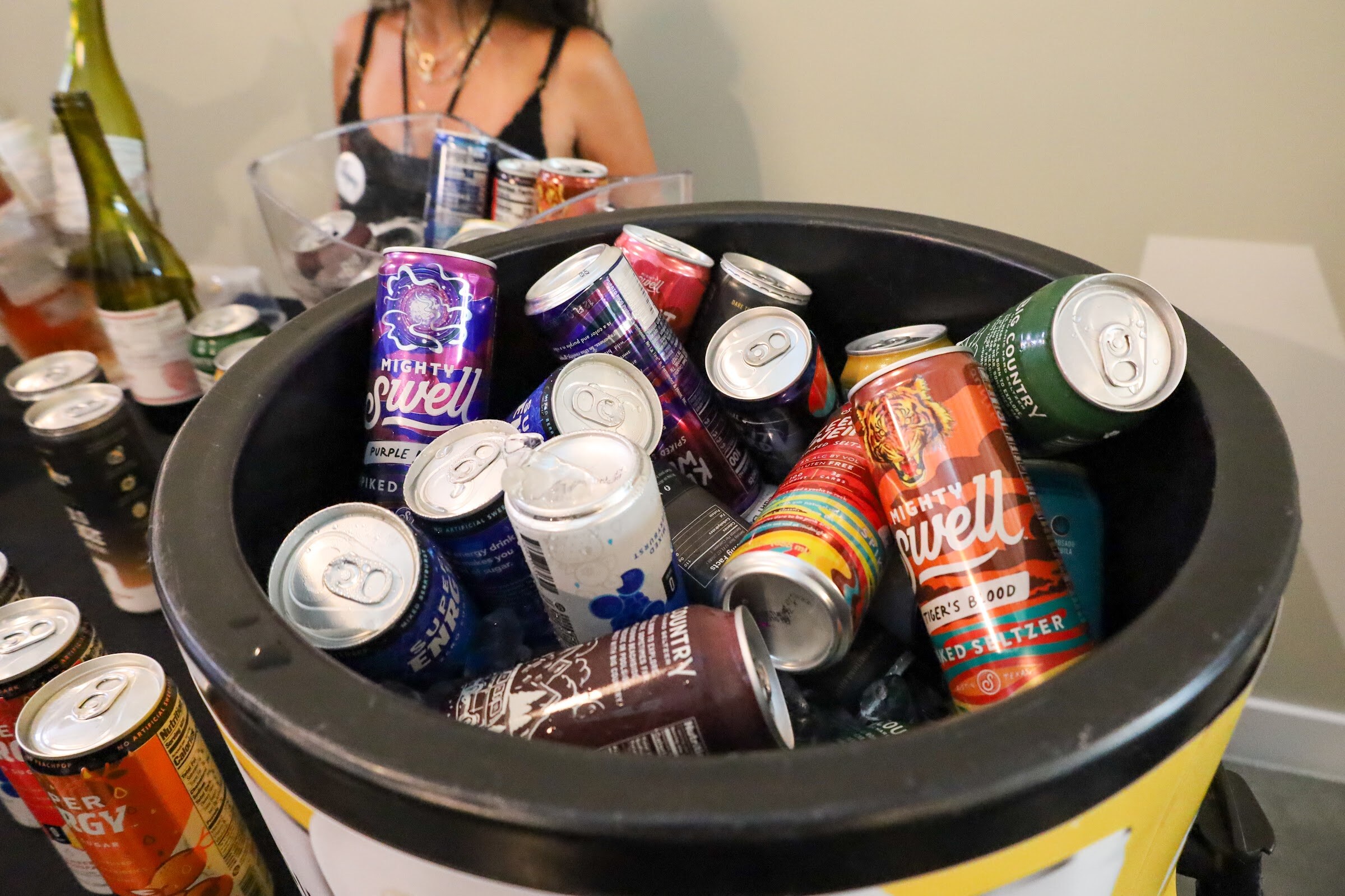 Close view into a black bucket full of cans, including Mighty Swell spiked seltzer in purple and Tiger's Blood flavors and Super Enrgy cans, with wine bottles and a woman in the background.