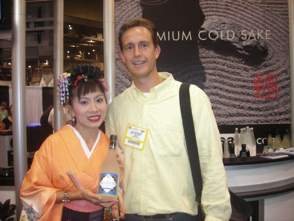 A woman in an orange kimono with a decorated hairpiece holds a bottle of Kigen sake beside a tall smiling man in a yellow shirt with a badge. A sign behind them reads Premium Cold Sake.