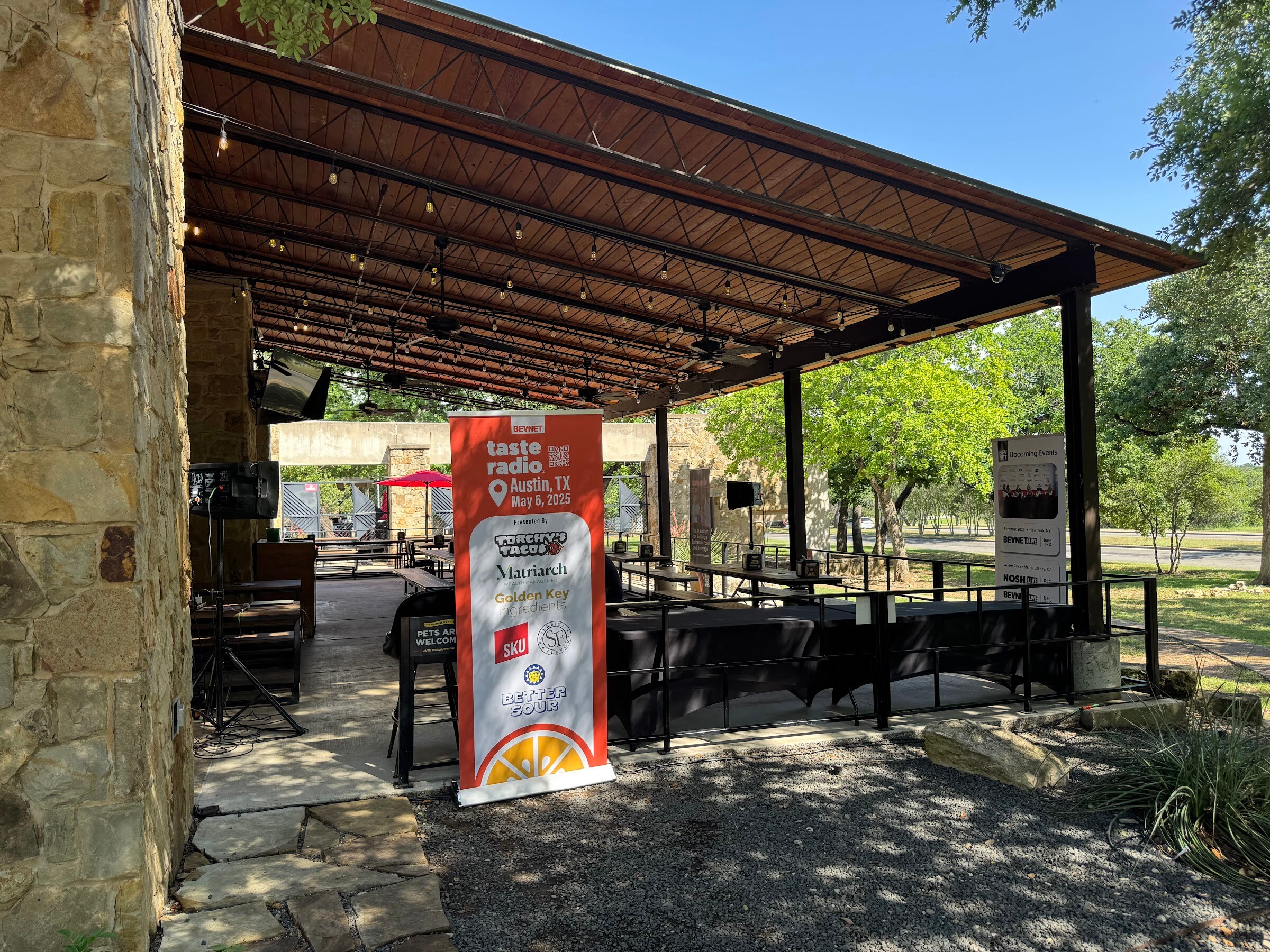 Orange Taste Radio banner at the end of a covered stone patio with picnic tables and trees beyond.