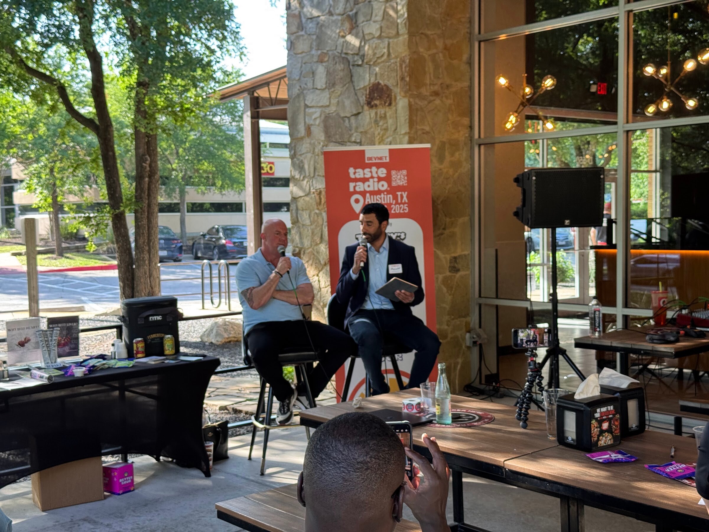 Two men seated on stools talking in front of a Taste Radio Austin banner, with a speaker and trees behind.