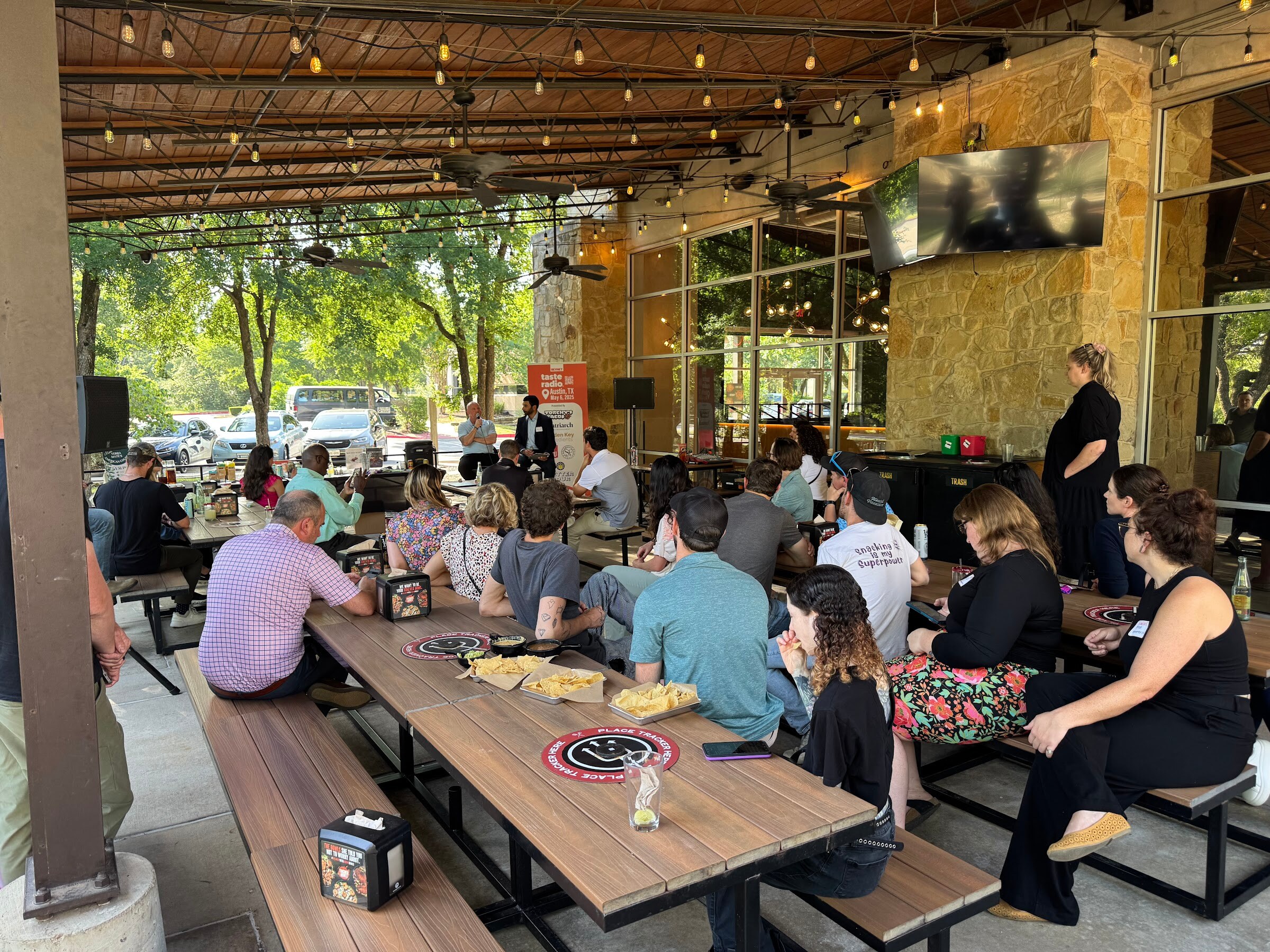 Seated audience at long wooden picnic tables under a covered patio, facing a small stage.