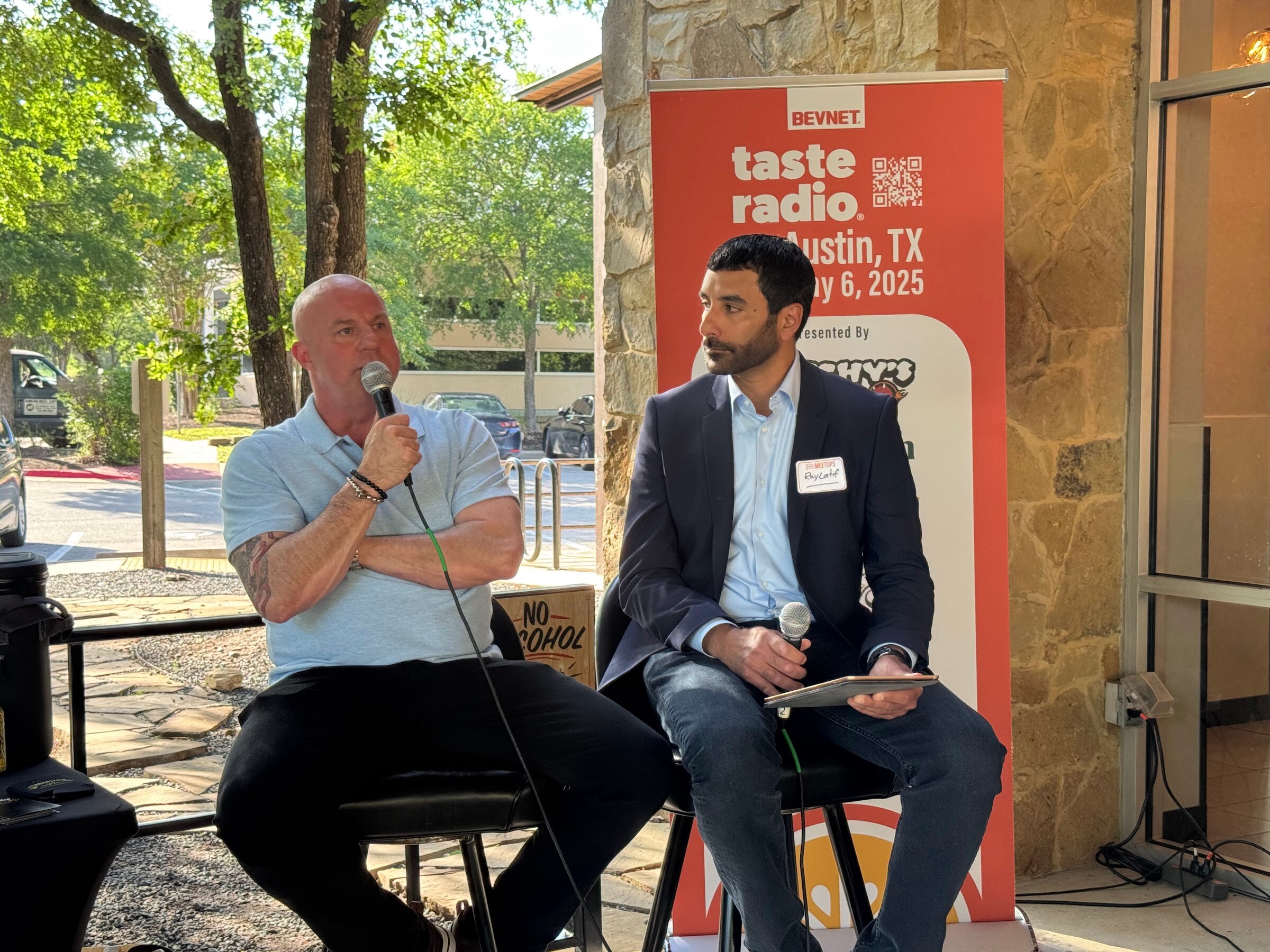 Man in a gray shirt with arms crossed seated beside a host holding a tablet, in front of the Taste Radio banner.