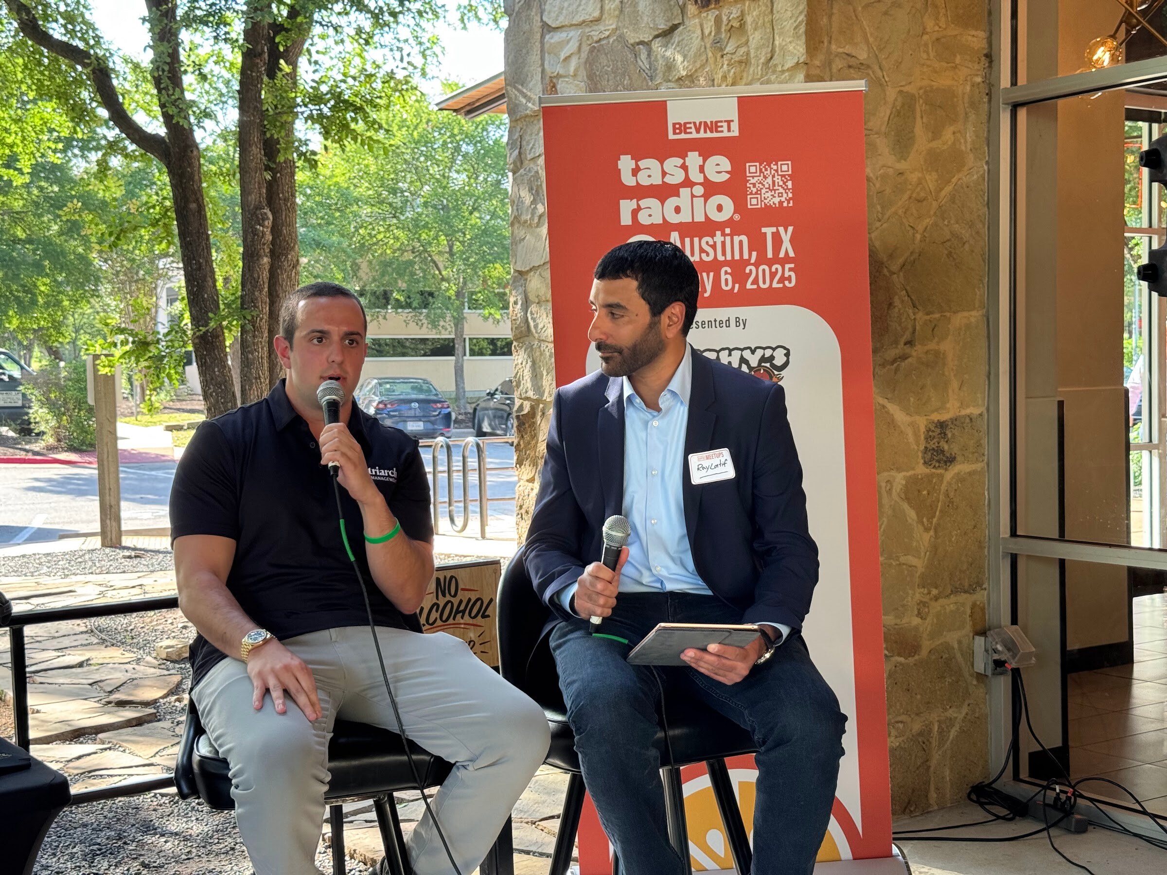 Two men seated on stools in front of the Taste Radio Austin banner, one holding a microphone and one a tablet.