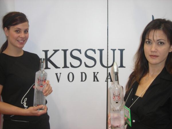 Two women in black each hold a bottle of Kissui Vodka in front of a white wall with the Kissui Vodka logo. The bottles carry a red and black mask design.