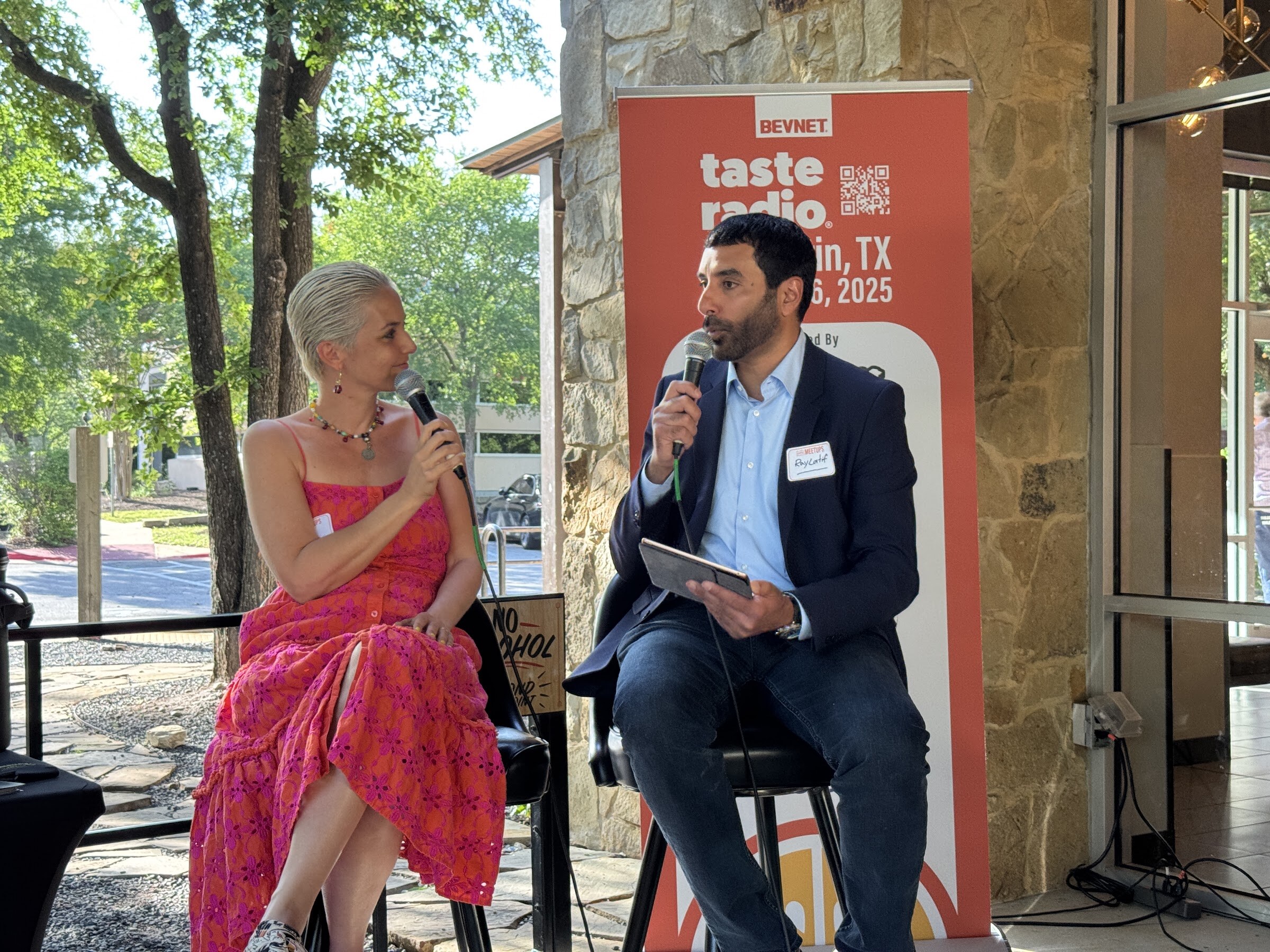 Woman in a red dress and a man in a navy suit seated on stools in front of the event banner.