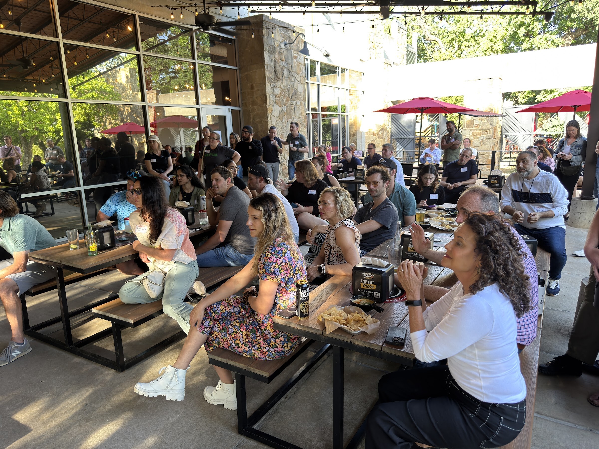 Crowd seated at picnic tables on a covered patio, watching a stage out of frame.