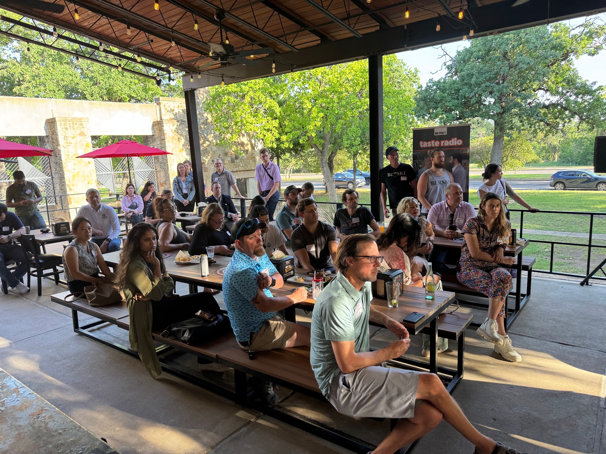Man in a green polo seated at a picnic table with a crowd behind him on the patio.