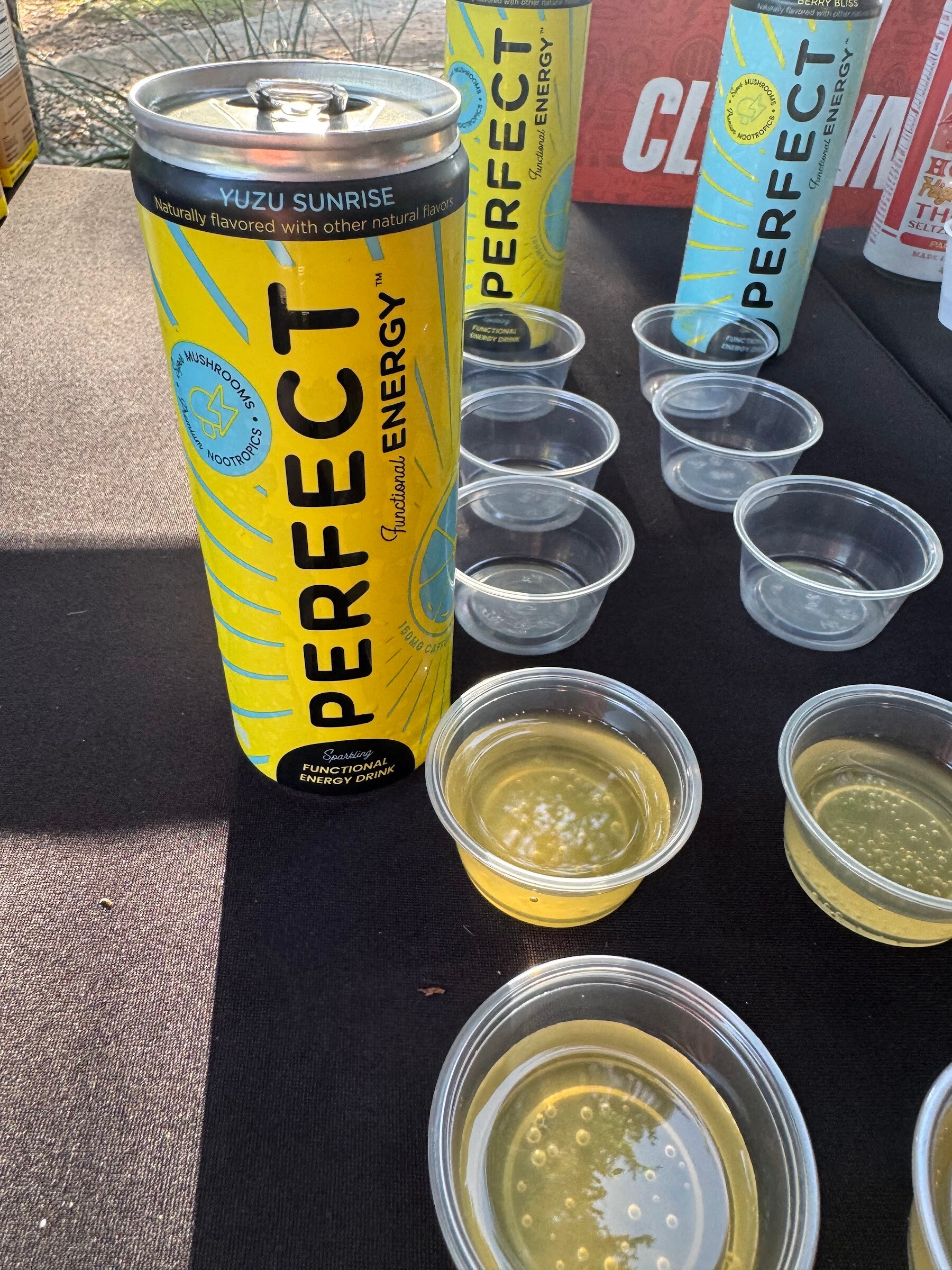 Tall yellow can of Perfect Energy Yuzu Sunrise next to small sample cups on a black tablecloth.