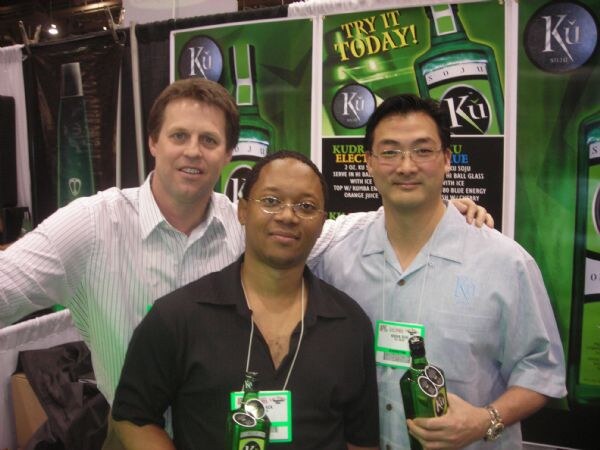 Three men pose close together in front of green Ku soju posters; one wears a white striped shirt, one a black shirt, one a light blue shirt and glasses, and two hold green bottles.