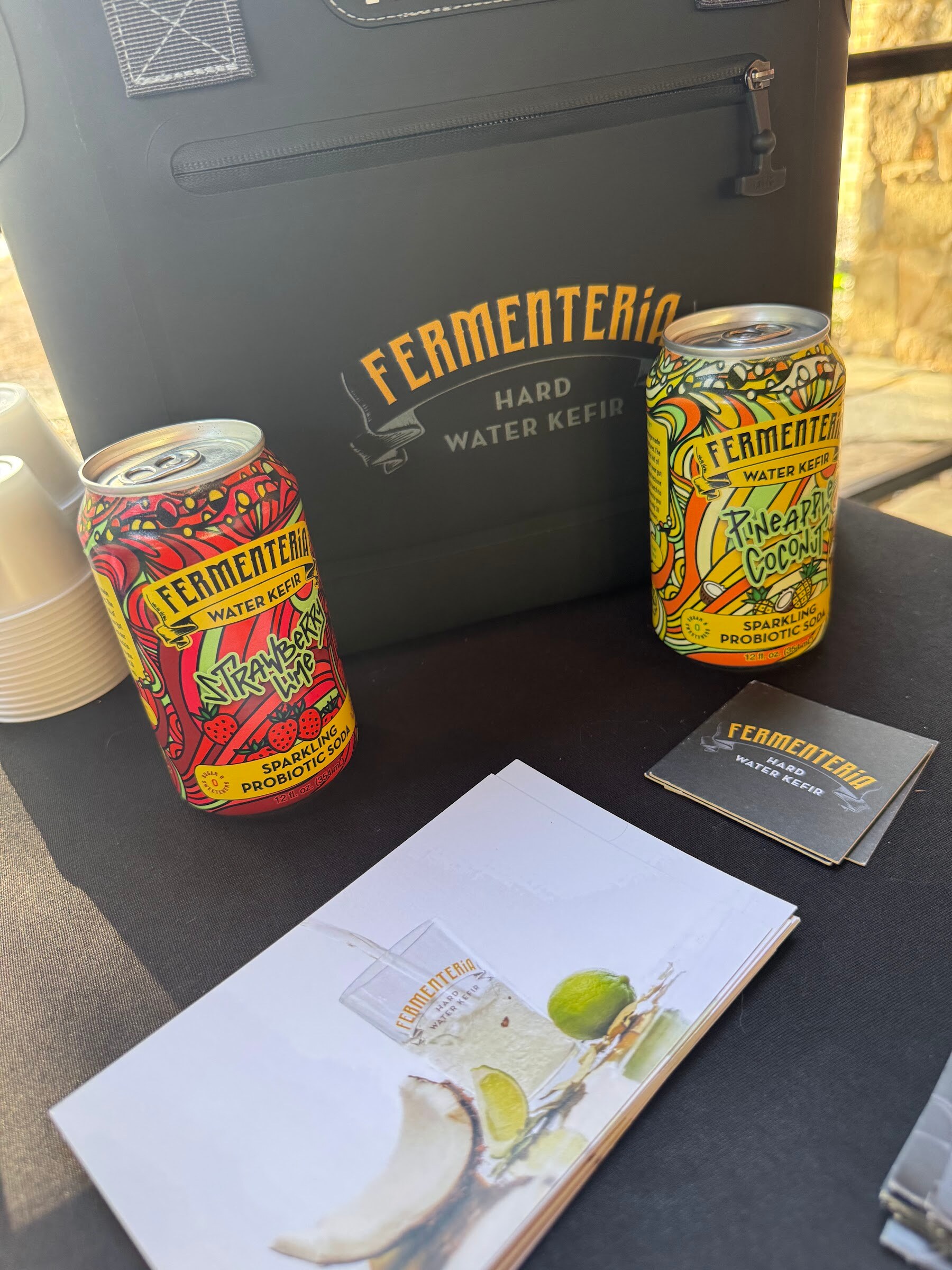 Two Fermentería cans on a table next to a black cooler and a flyer.