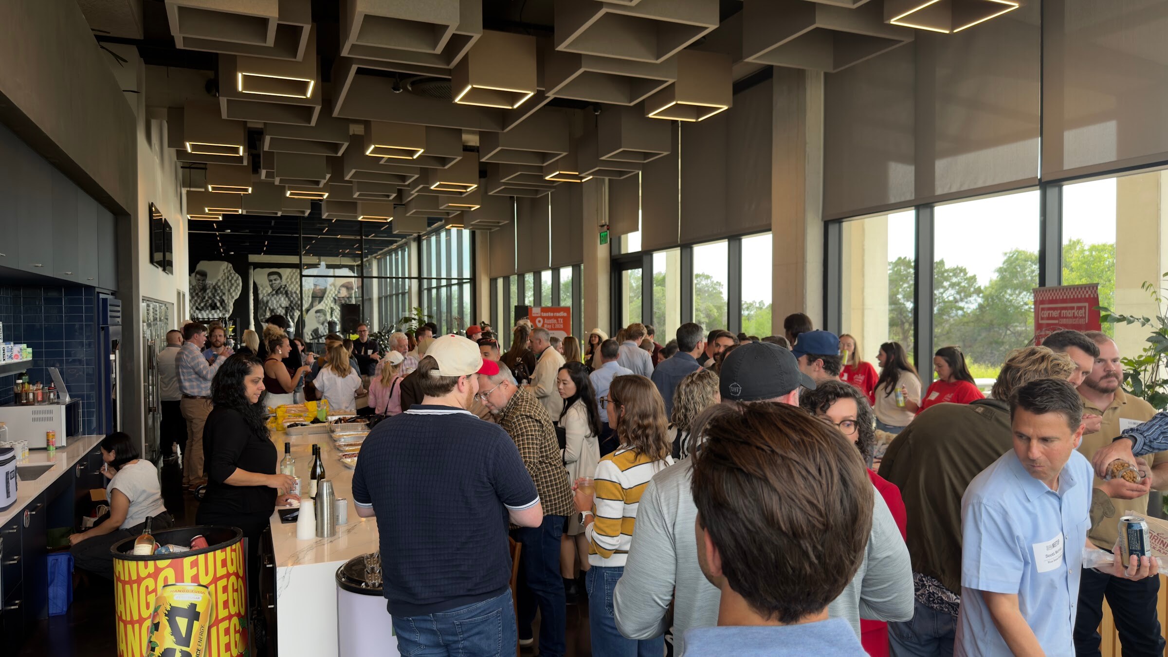 Wide view of attendees mingling along a long counter under a coffered ceiling with floor-to-ceiling windows.