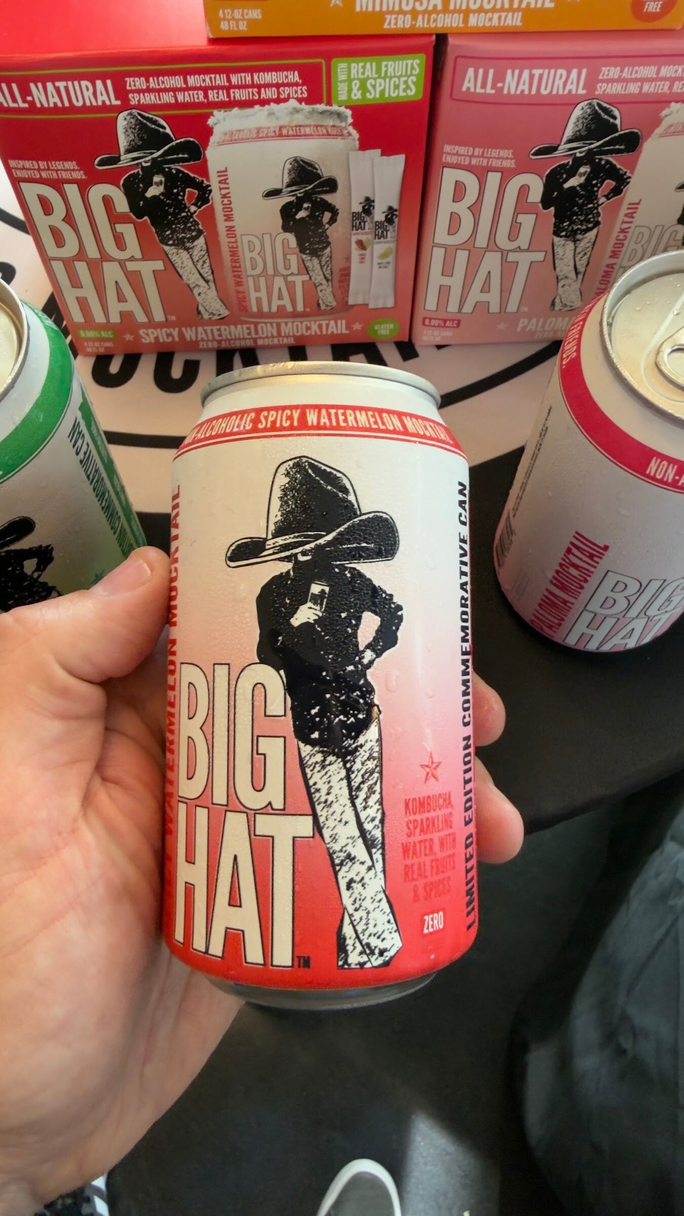 Hand holding a white and red Big Hat can in front of stacked Big Hat cases.