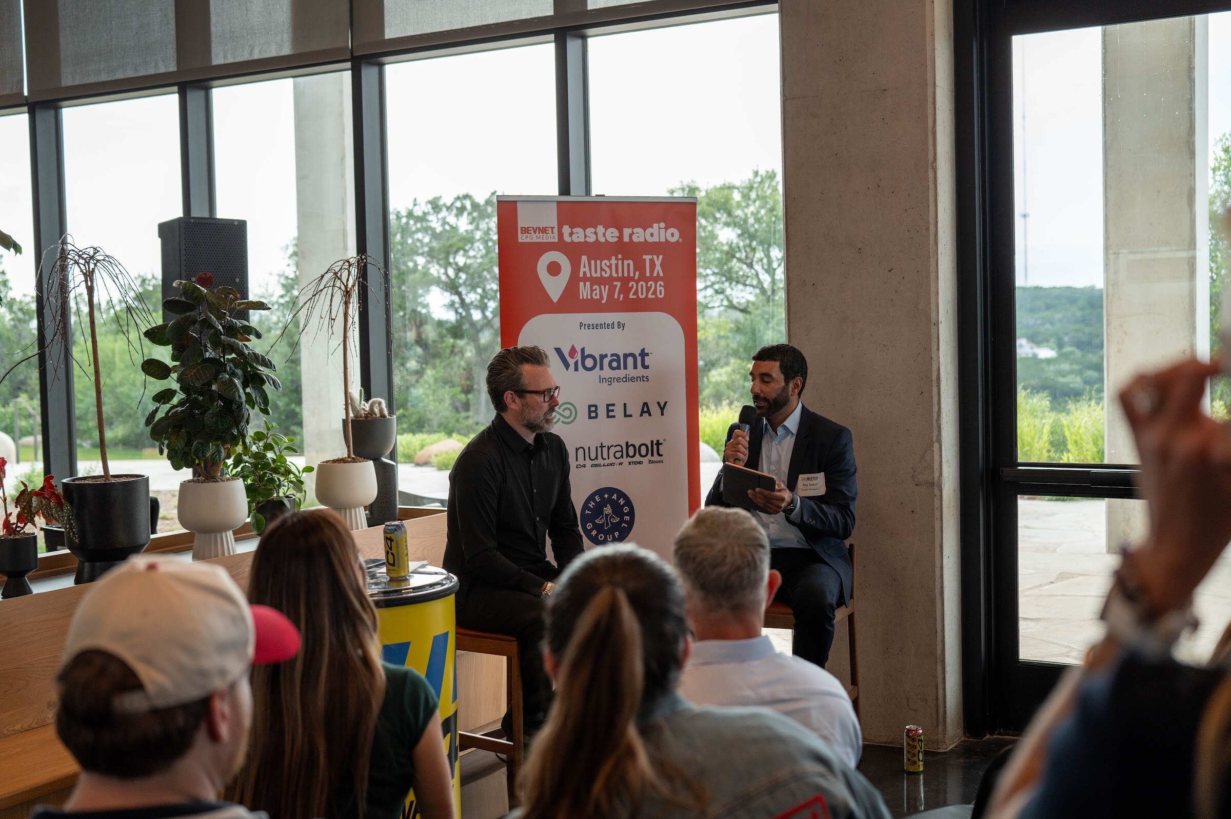 Host in a navy suit with a microphone faces a guest in black on stools beside the Taste Radio Austin May 7, 2026 banner, audience in the foreground.