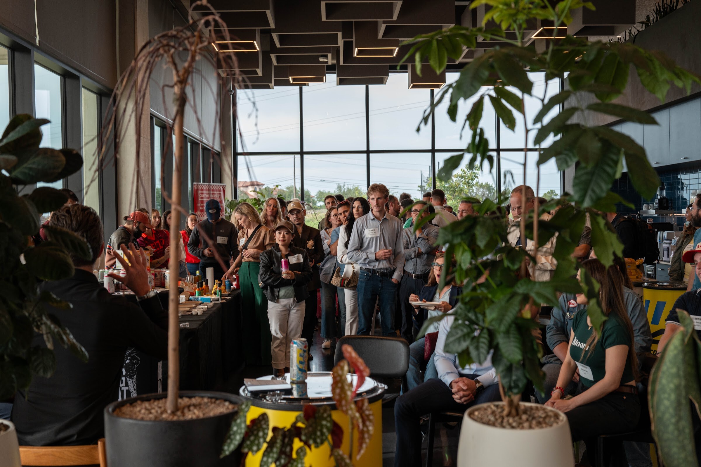 Crowd standing in a bright room with tall windows, seen between potted plants.