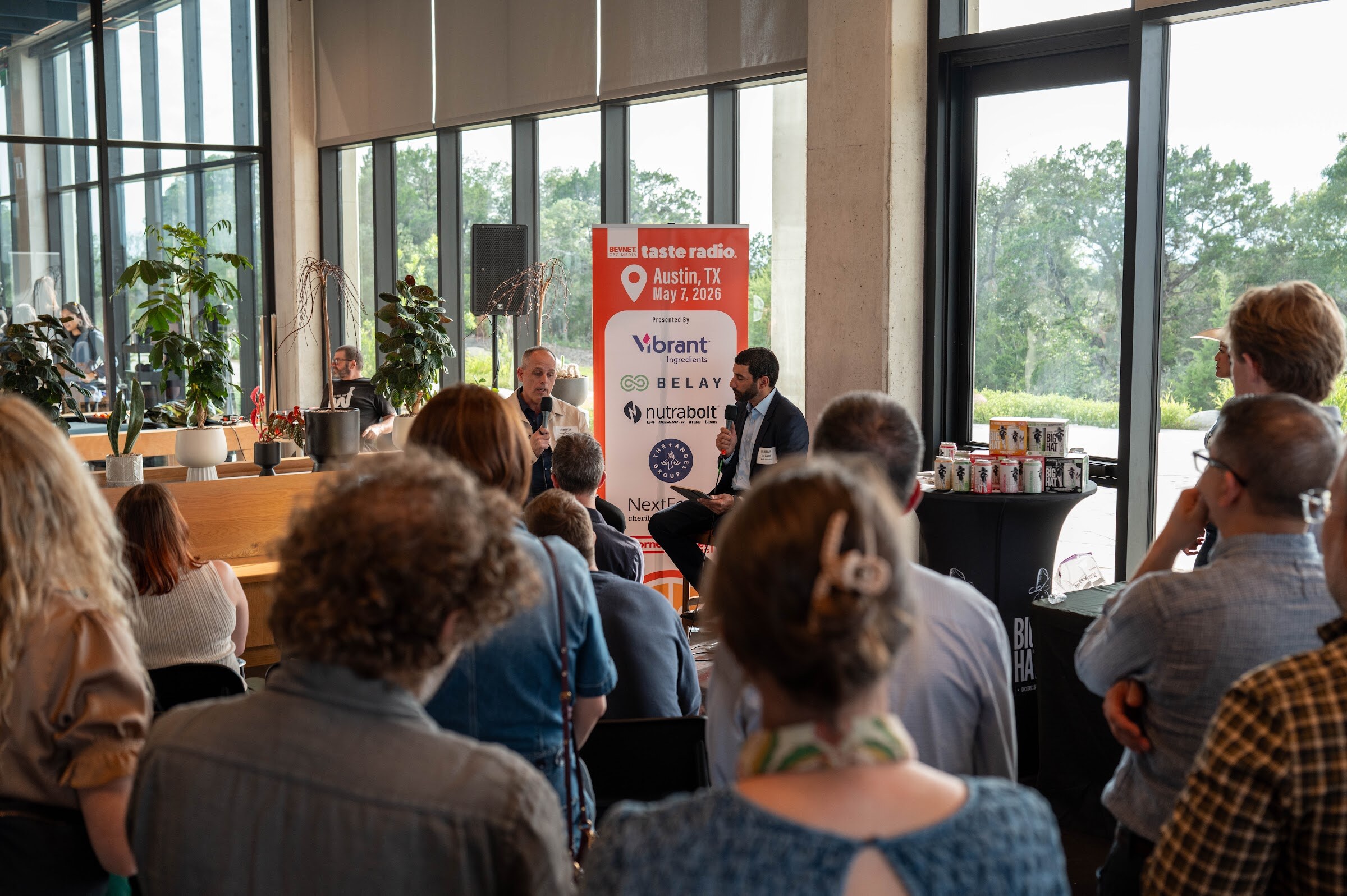 Seated audience in front of two people on stools beside the Taste Radio banner and windows.