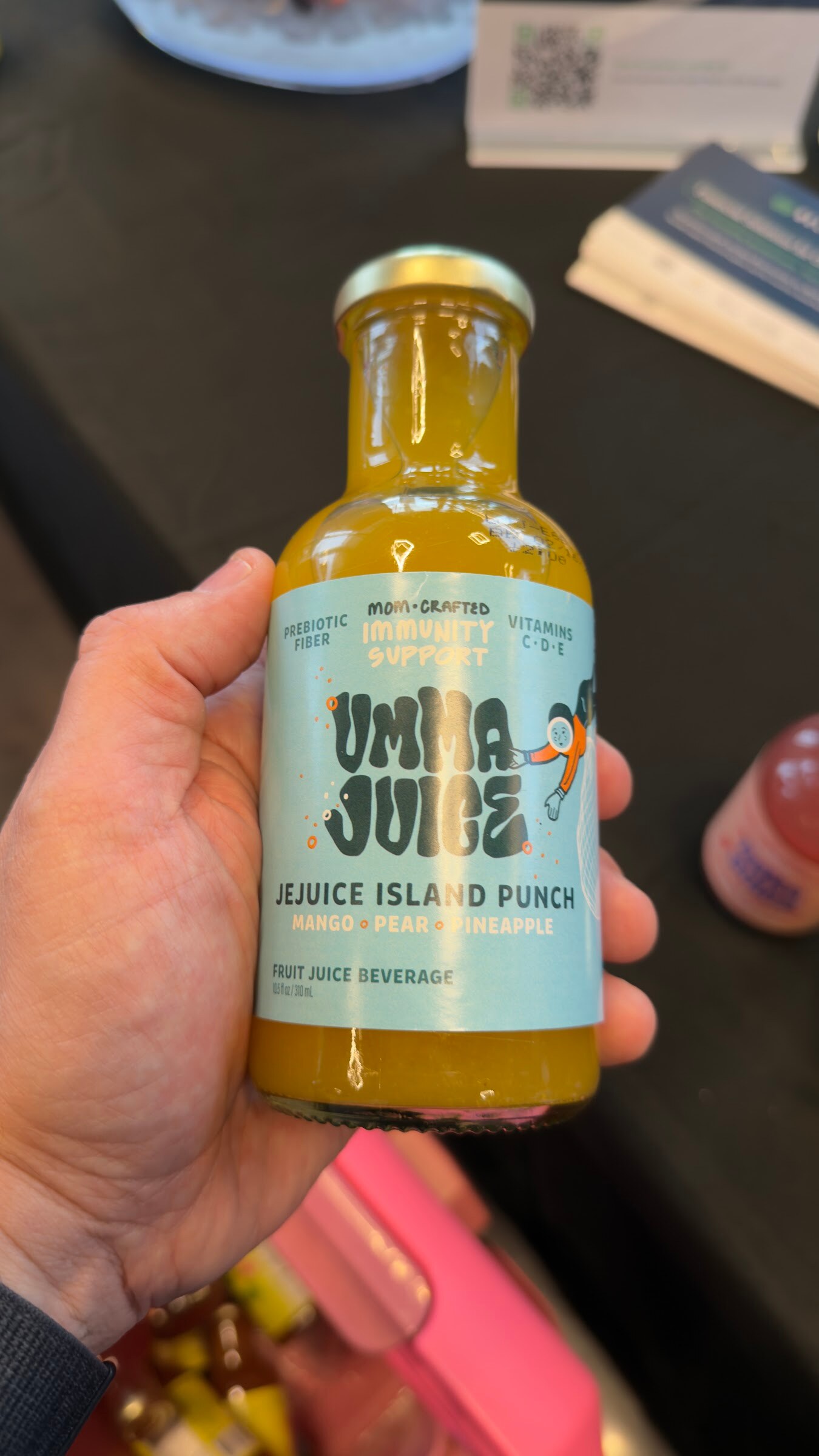Hand holding a glass bottle of Umma Juice Jejuice Island Punch over a sampling table.