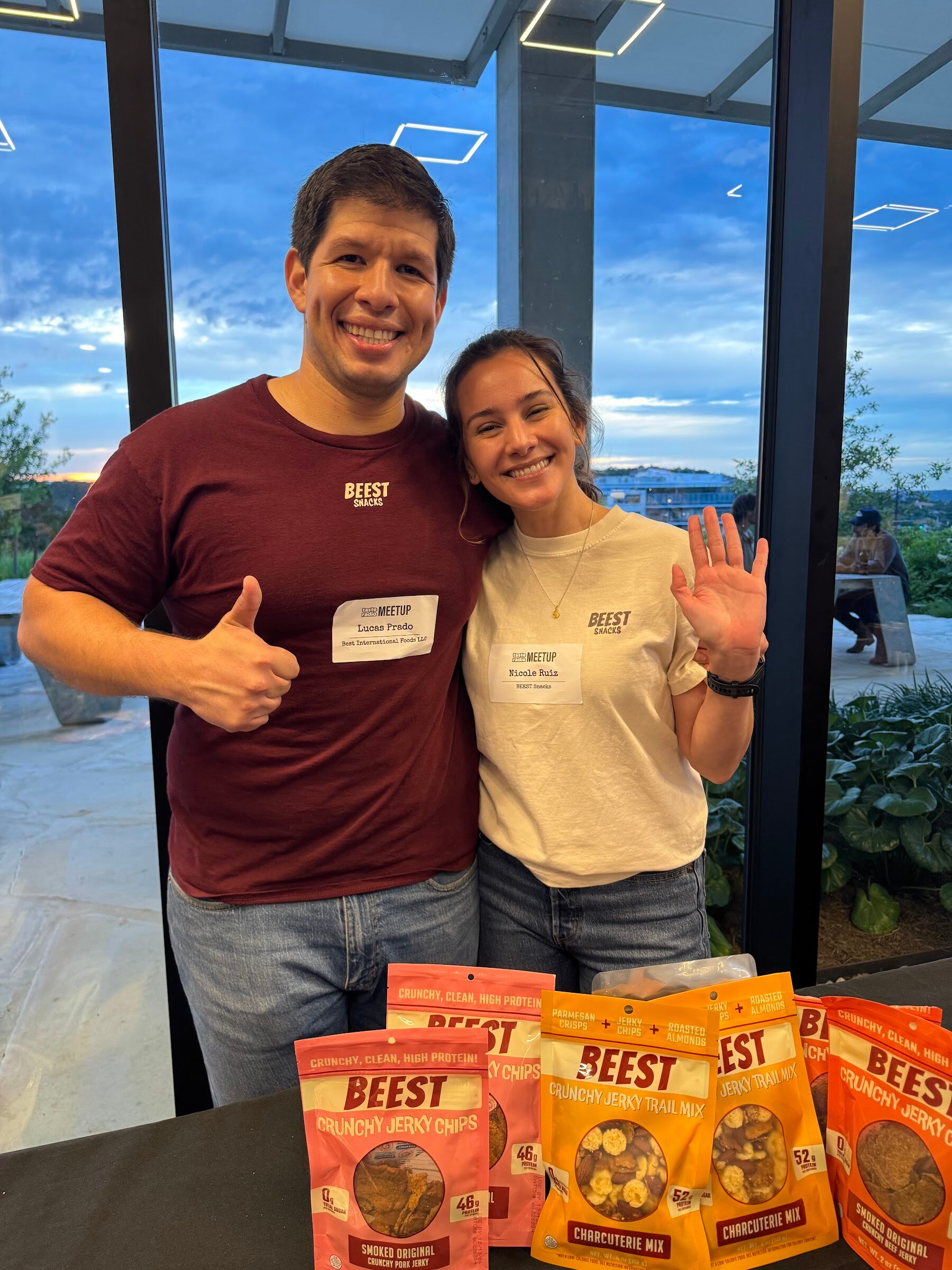 Smiling man giving a thumbs-up and a woman waving behind bags of Beest crunchy jerky chips and trail mix.