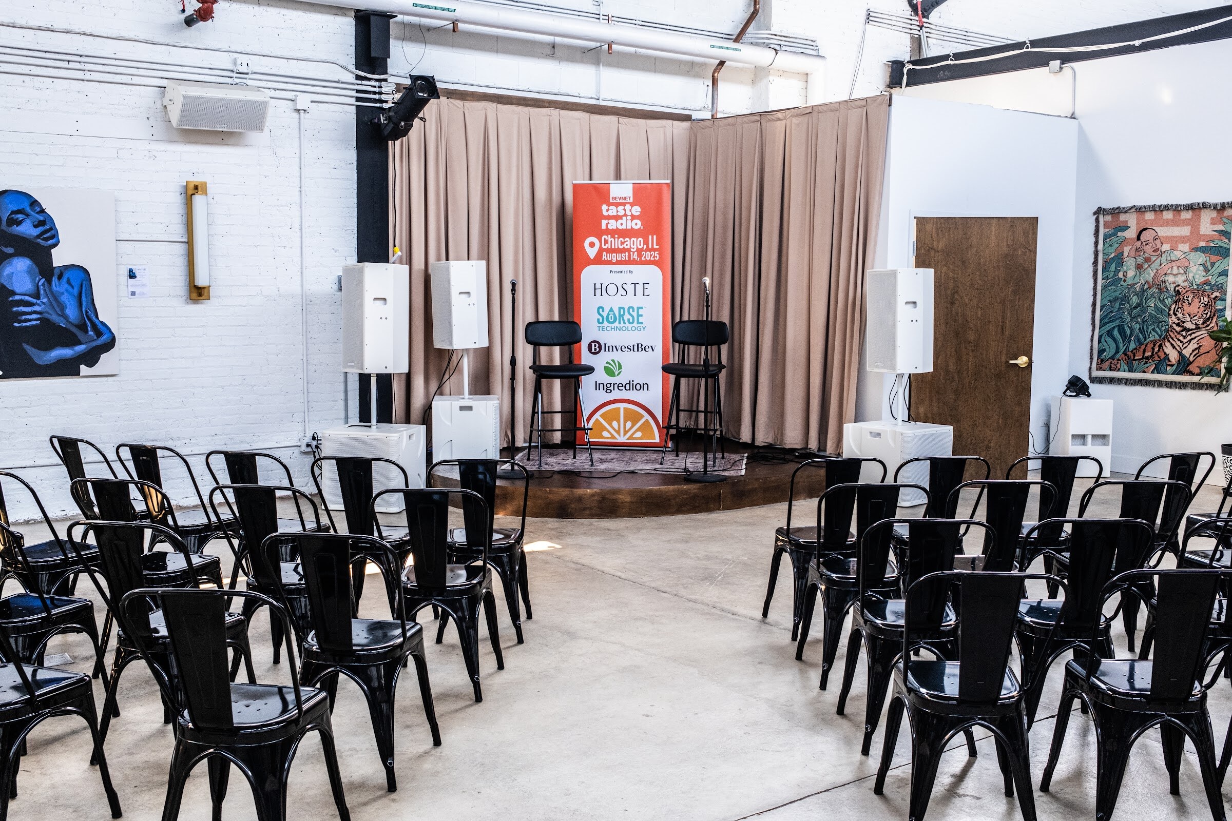 Empty black metal chairs facing a small stage with a Taste Radio banner and beige curtains.
