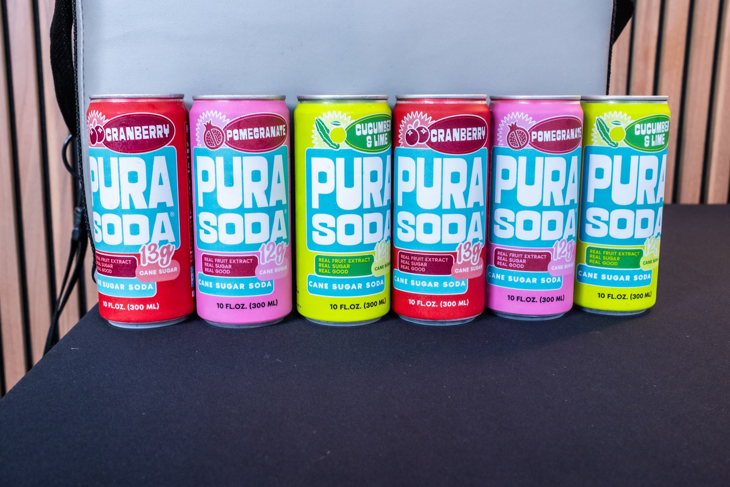 Row of Pura Soda cans in red, pink and green on a black tablecloth.