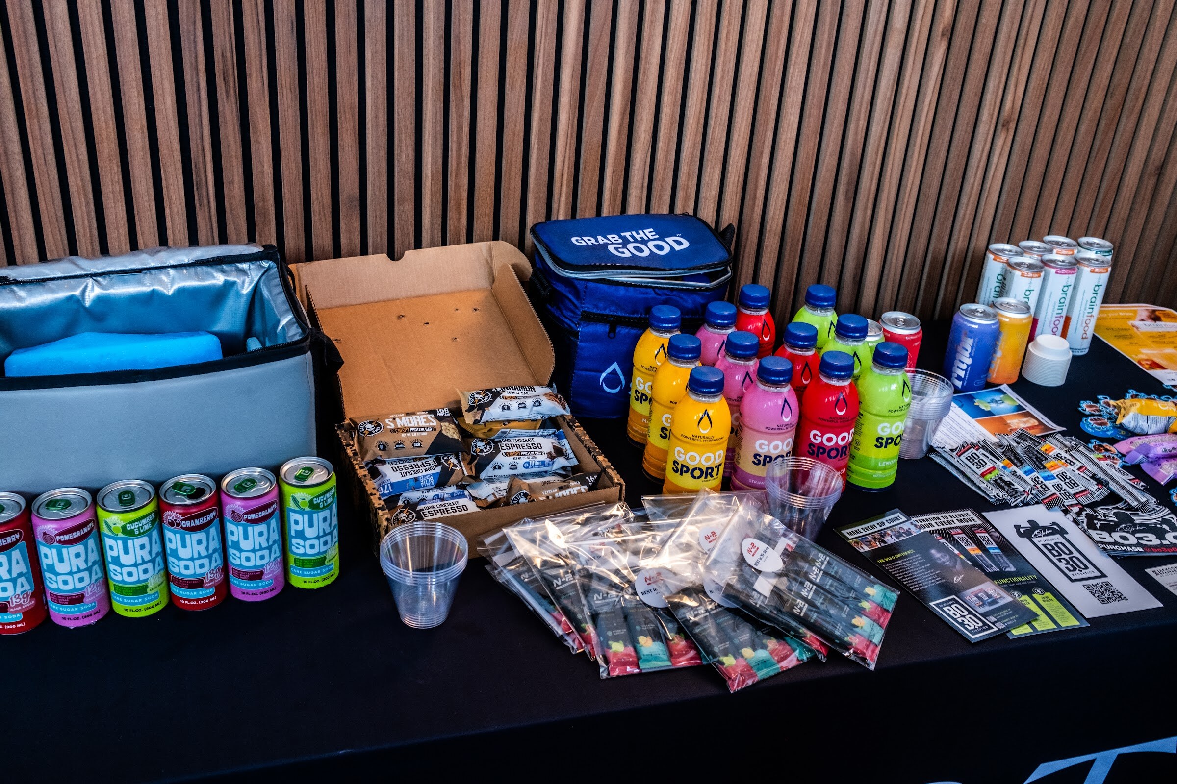 Table covered in canned drinks, snack bars and stickers, with a black cloth printed with Hoste.