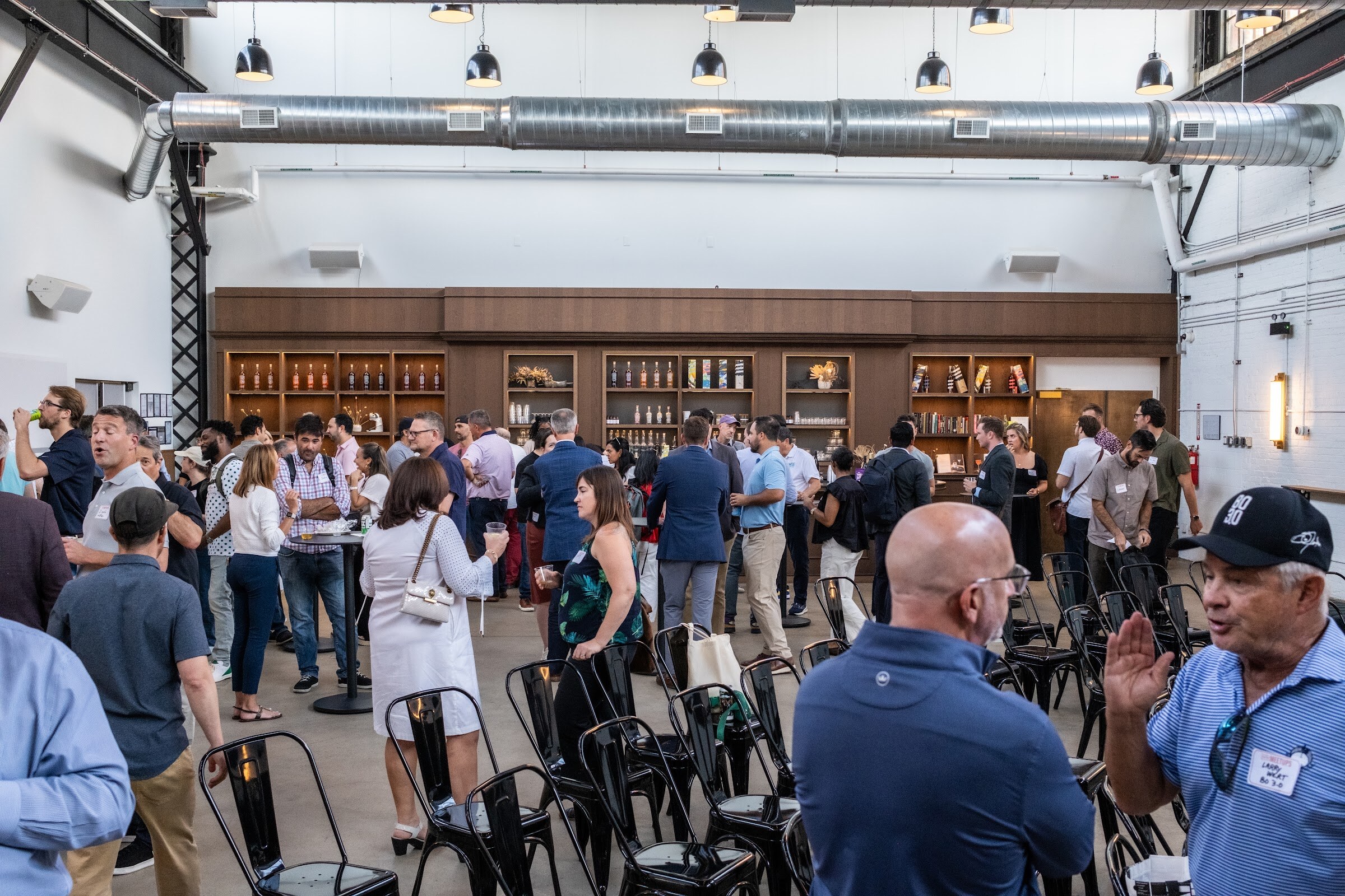 Crowd of attendees talking in a large brick-and-white room with a bar behind them.