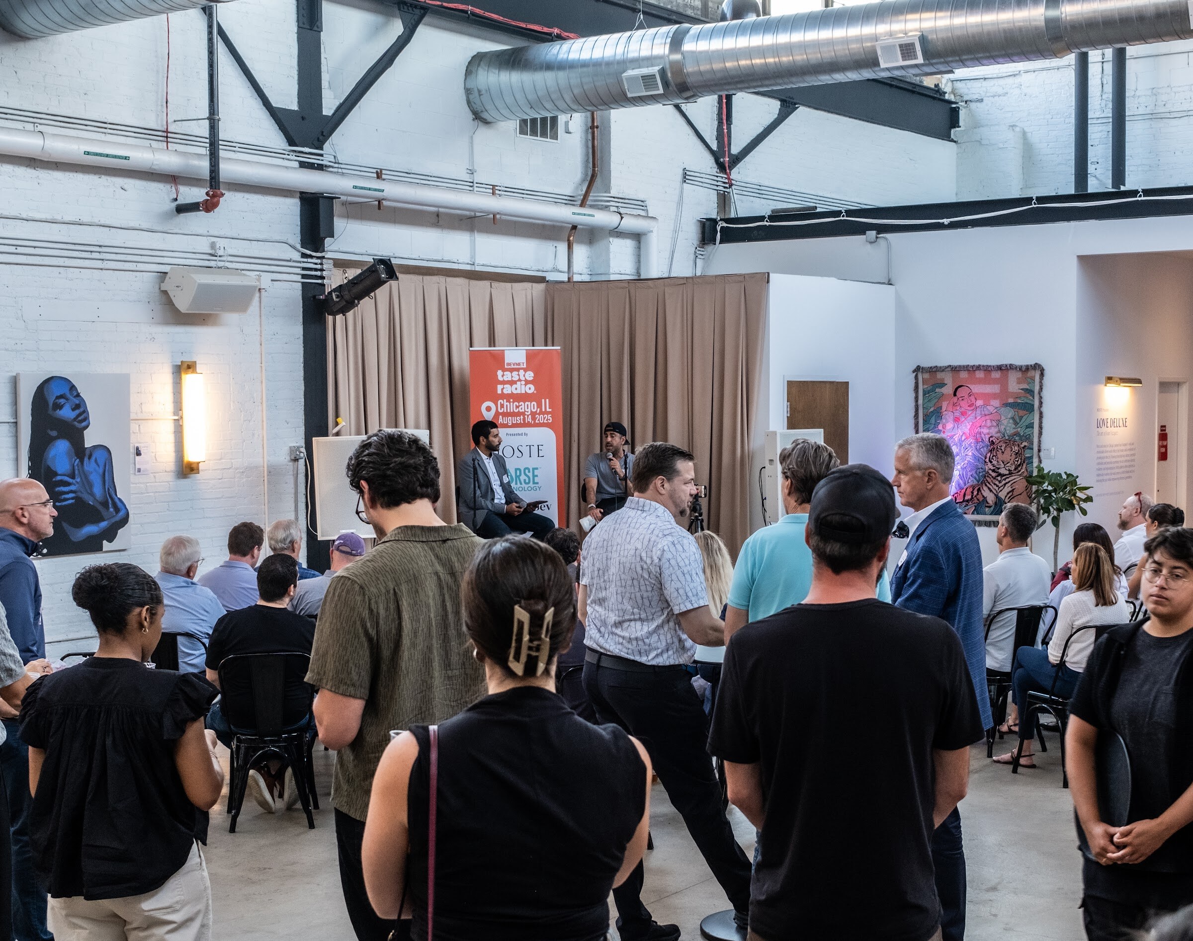 Crowd of attendees standing and seated facing a stage with an orange banner and beige curtains.