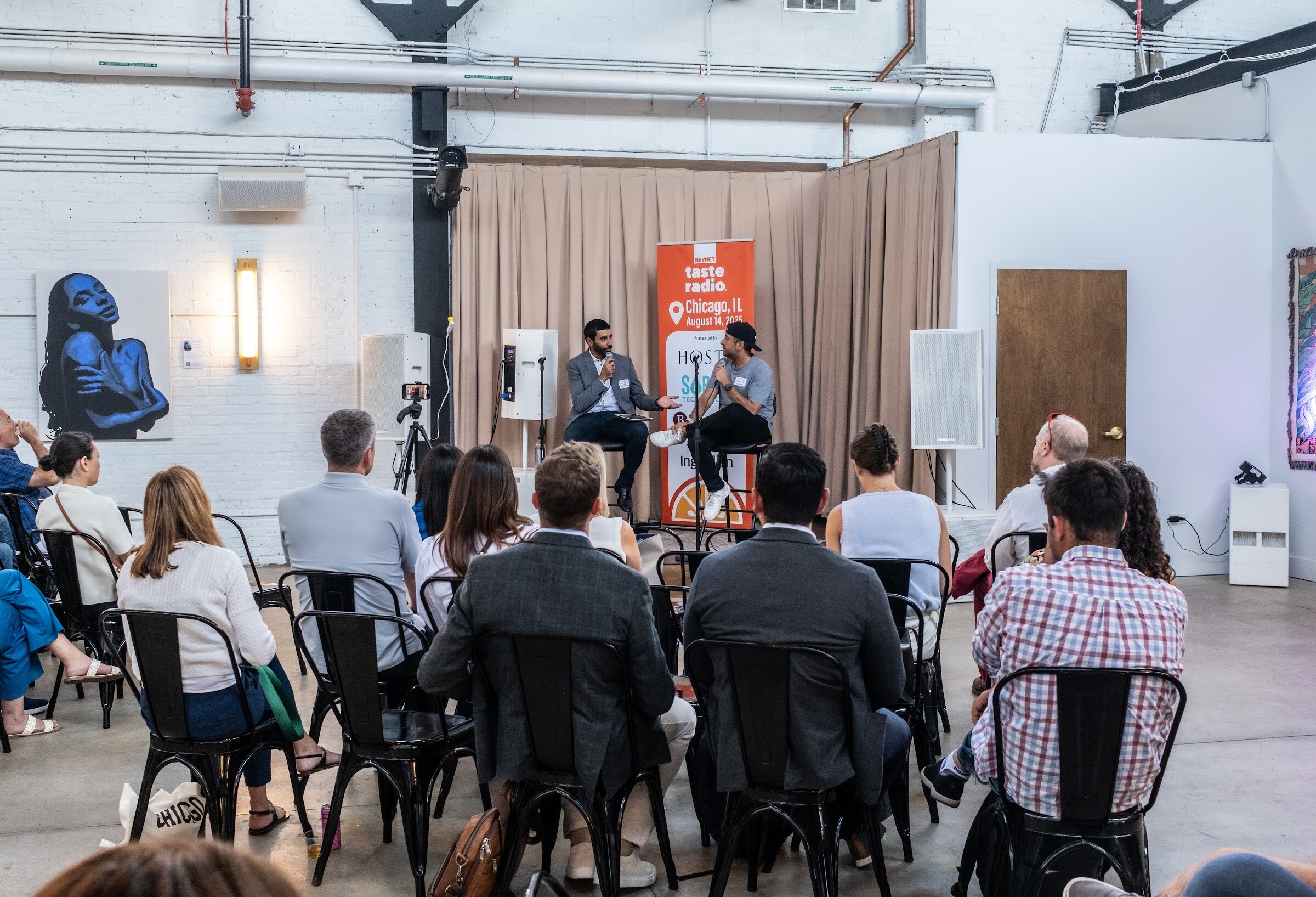 Seated and standing audience watching two people talking on a small stage in the distance.